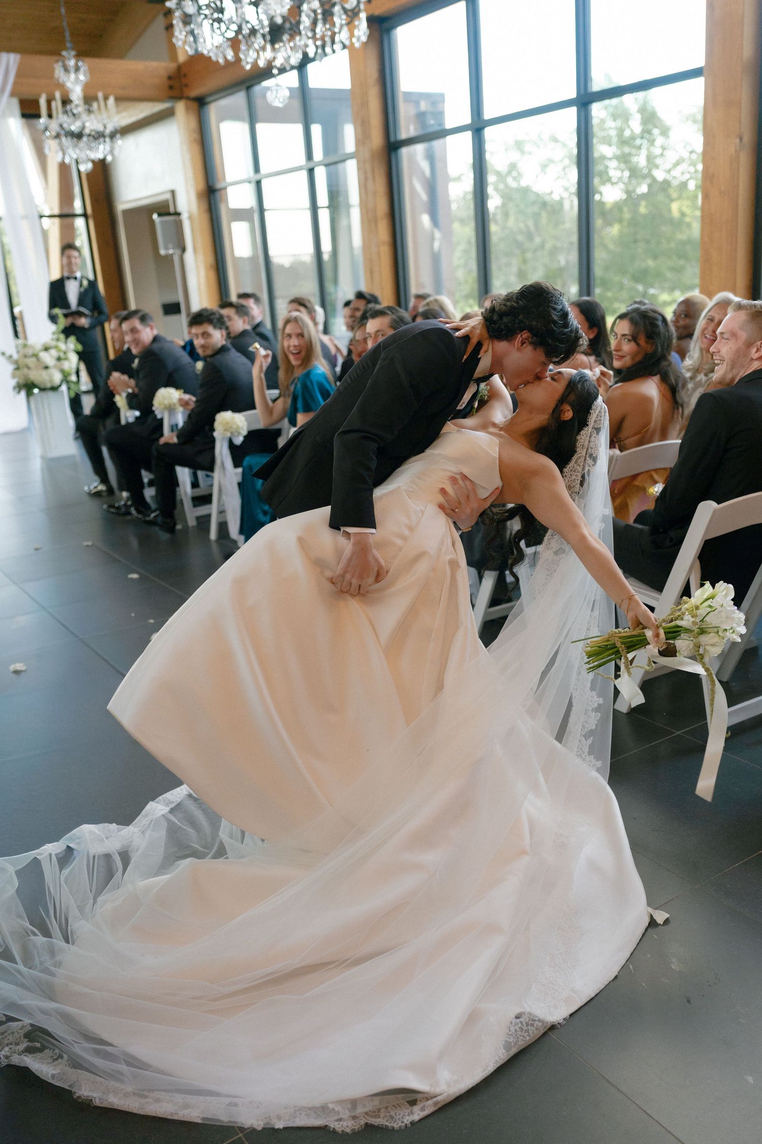 Groom dips bride for a romantic kiss during wedding ceremony in elegant venue with large windows.
