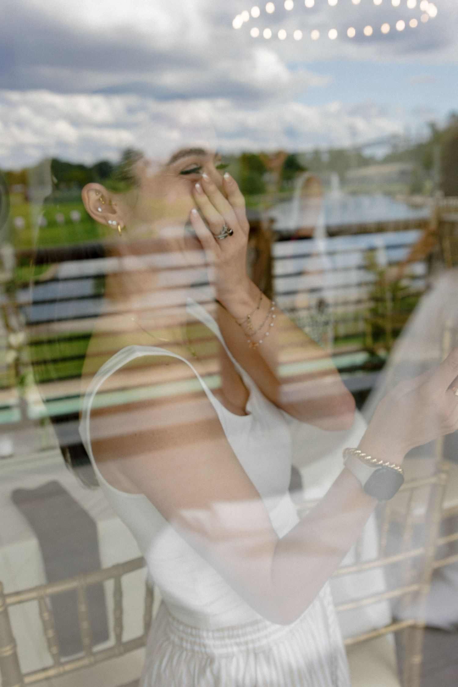 Bride laughing and covering her mouth, photographed through a window with outdoor reflection.