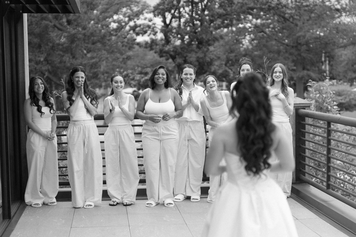Black and white photo of a bride revealing her wedding dress to smiling bridesmaids on an outdoor deck.