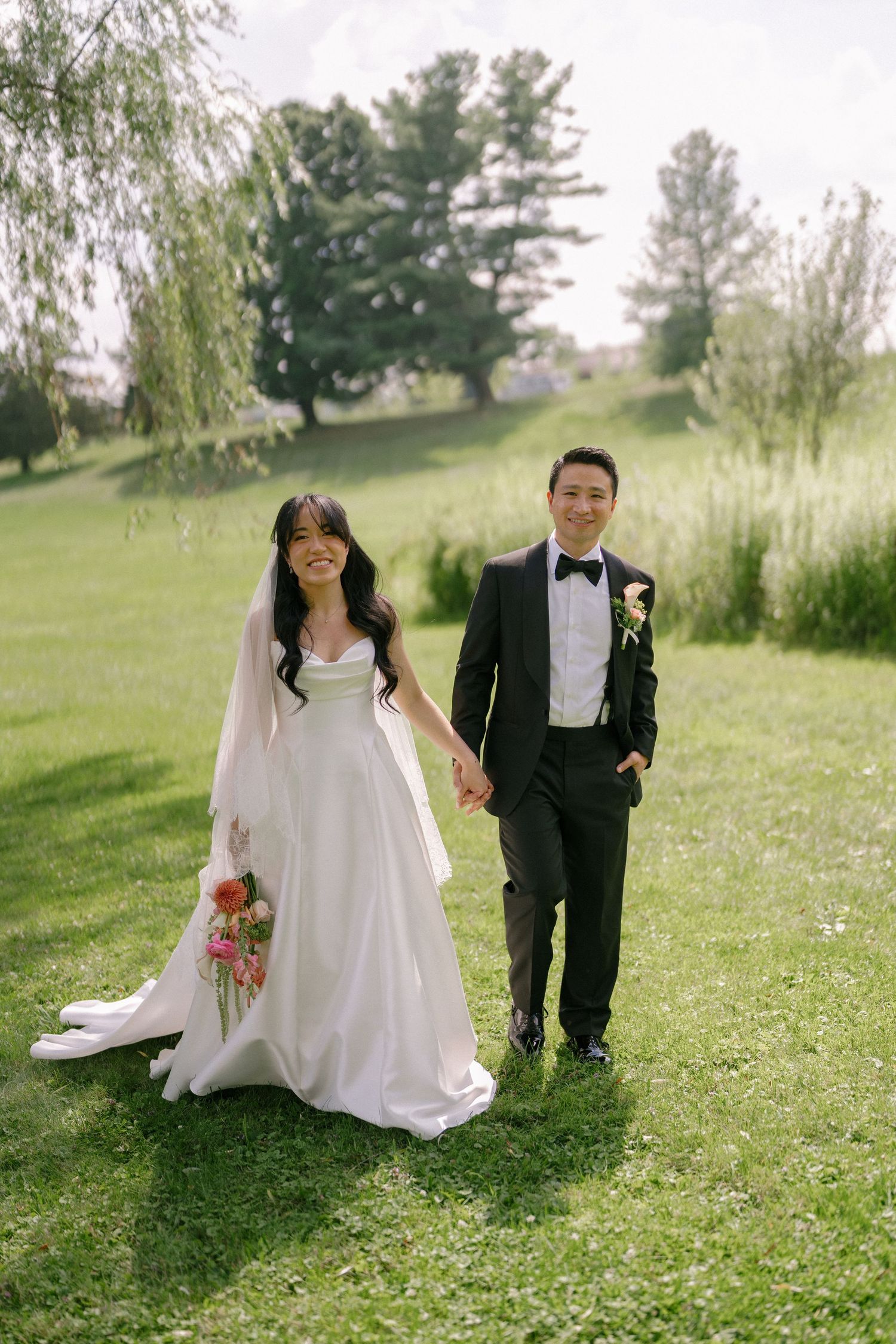 Bride in white gown with long veil and groom in black tuxedo holding hands on lush green lawn.
