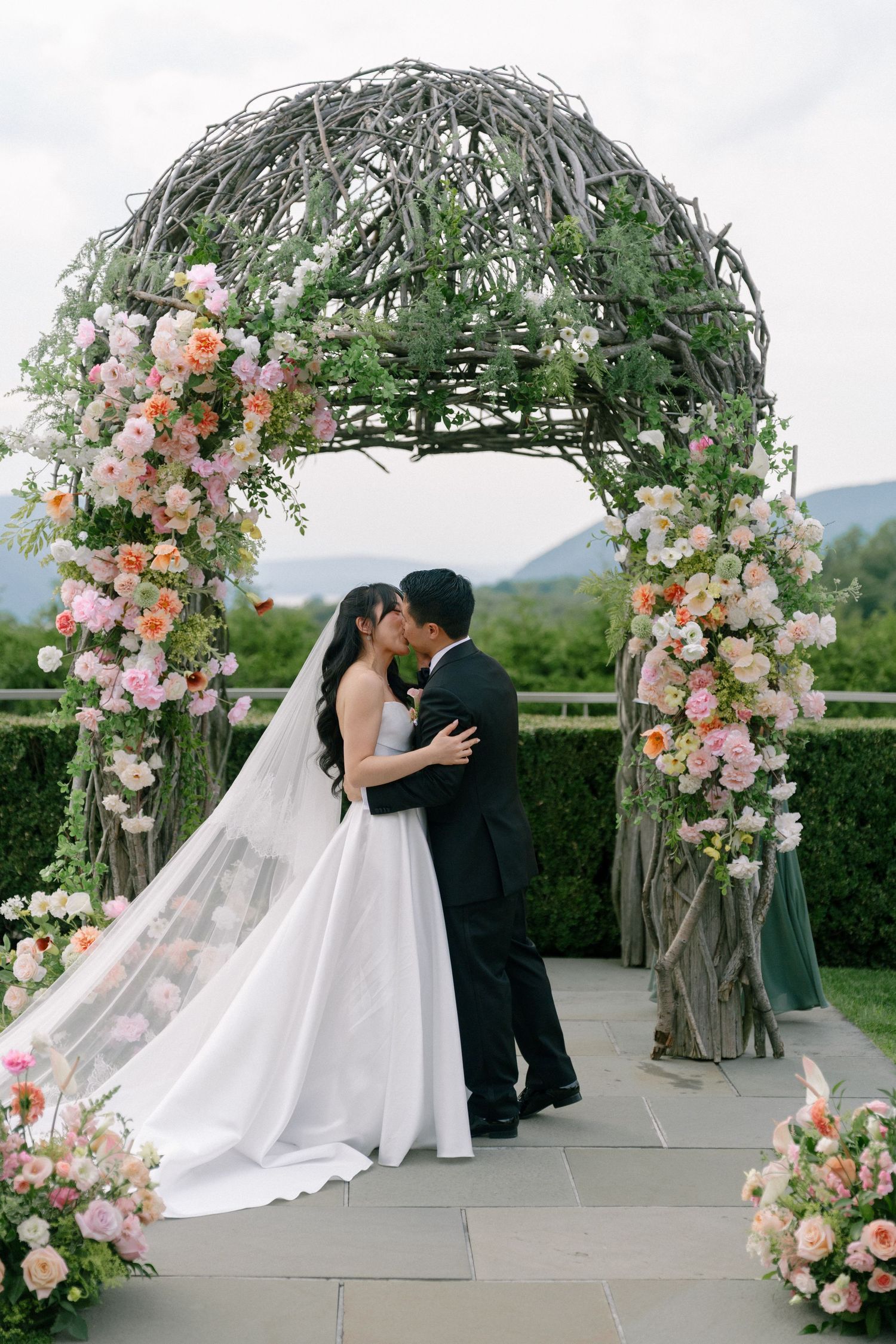 Bride and groom share first kiss under a floral arch adorned with pink and peach blooms at an outdoor wedding ceremony.