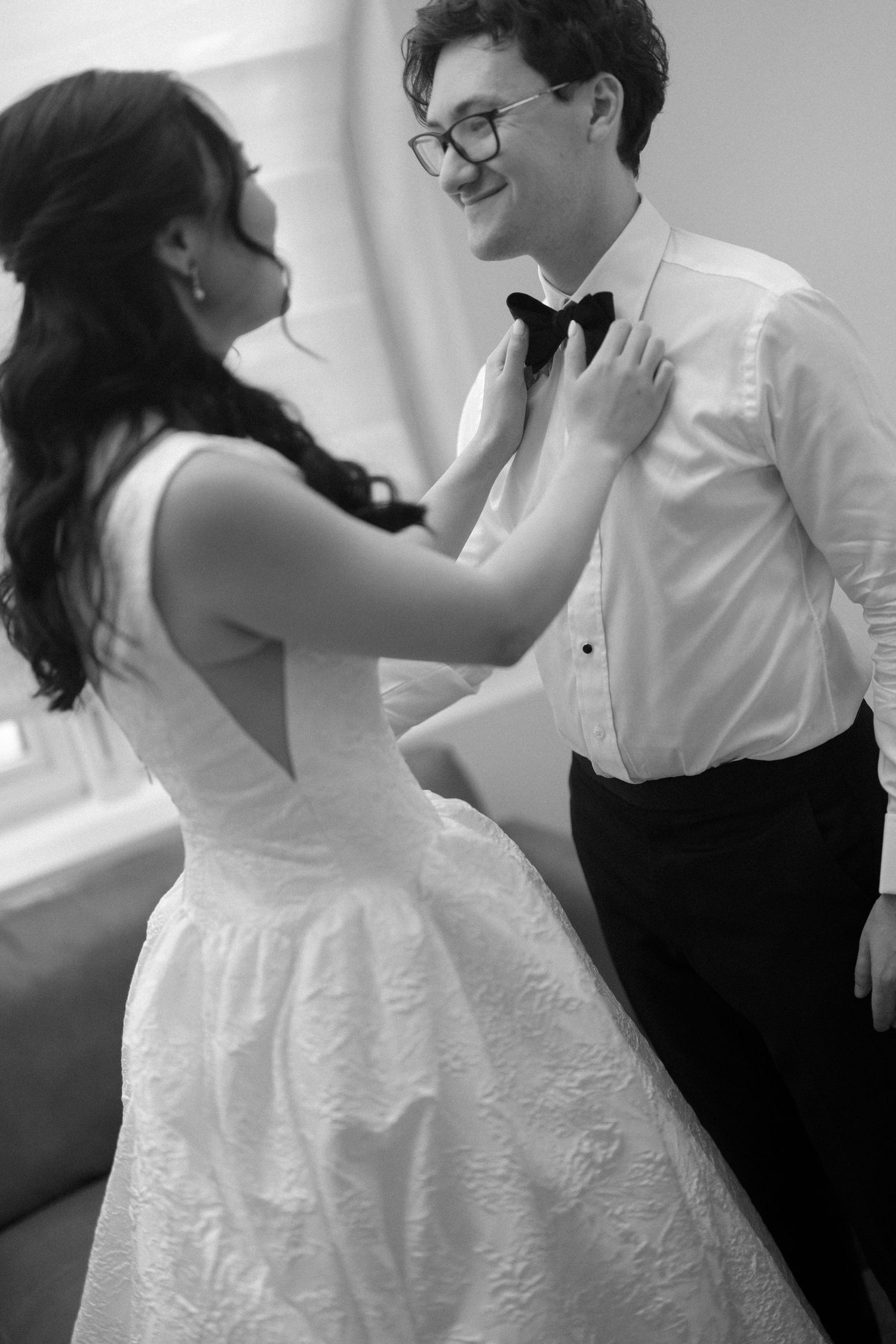 Black and white photo of a bride in lace gown adjusting groom's bow tie, both smiling at each other.