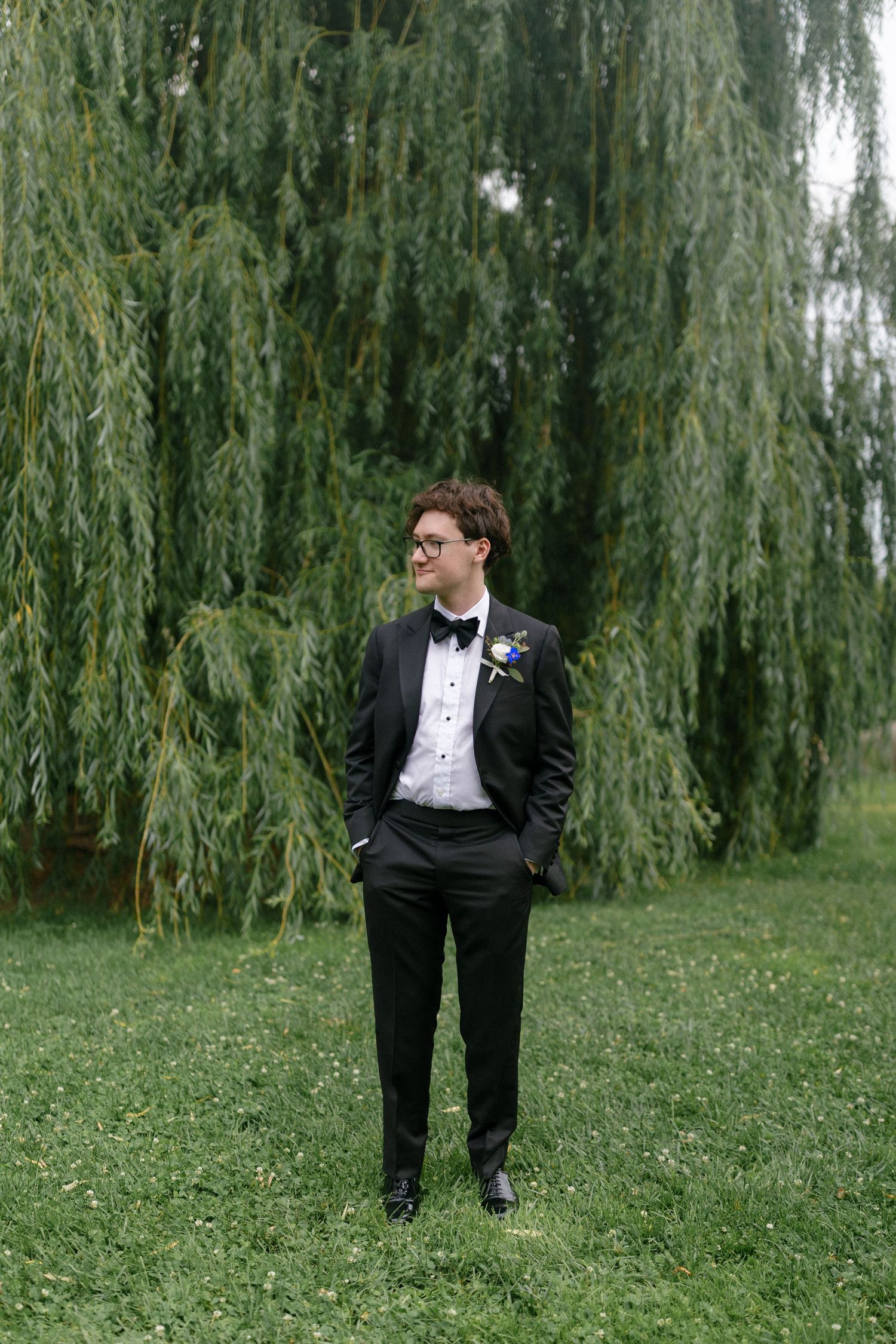 Groom in black tuxedo with boutonniere stands confidently before a weeping willow tree on green grass.