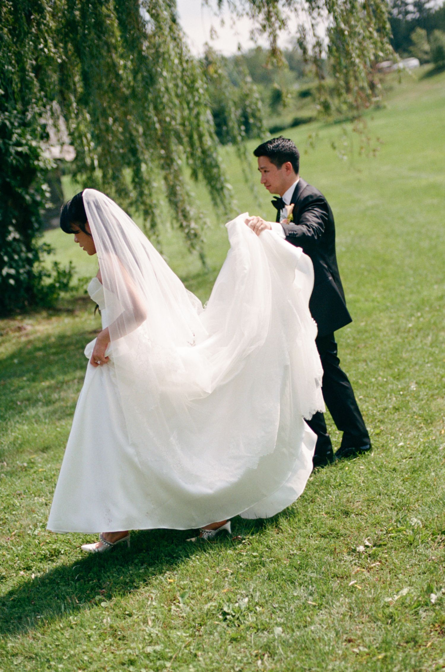 Groom in black tuxedo carrying bride's white wedding gown train on green lawn near weeping willow tree.