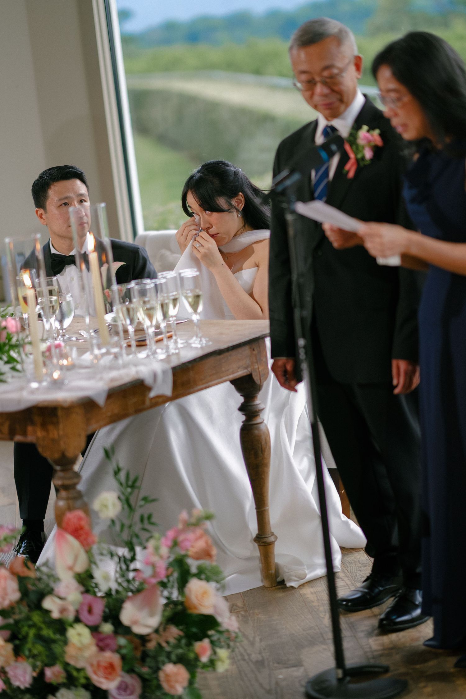 Emotional bride wiping tears at sweetheart table while parents deliver heartfelt speech at elegant wedding reception.