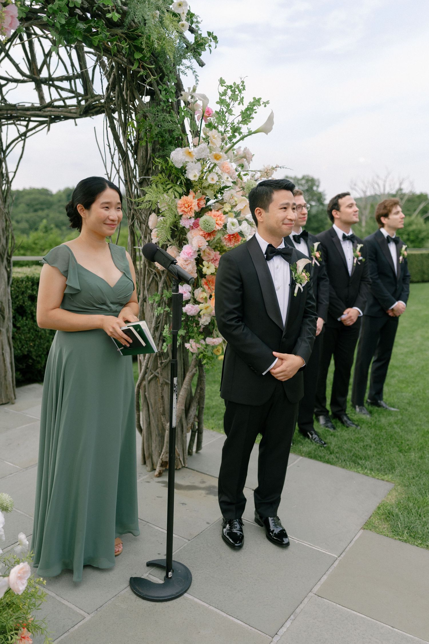 Groom in black tuxedo stands at outdoor wedding altar with floral arch and wedding party.
