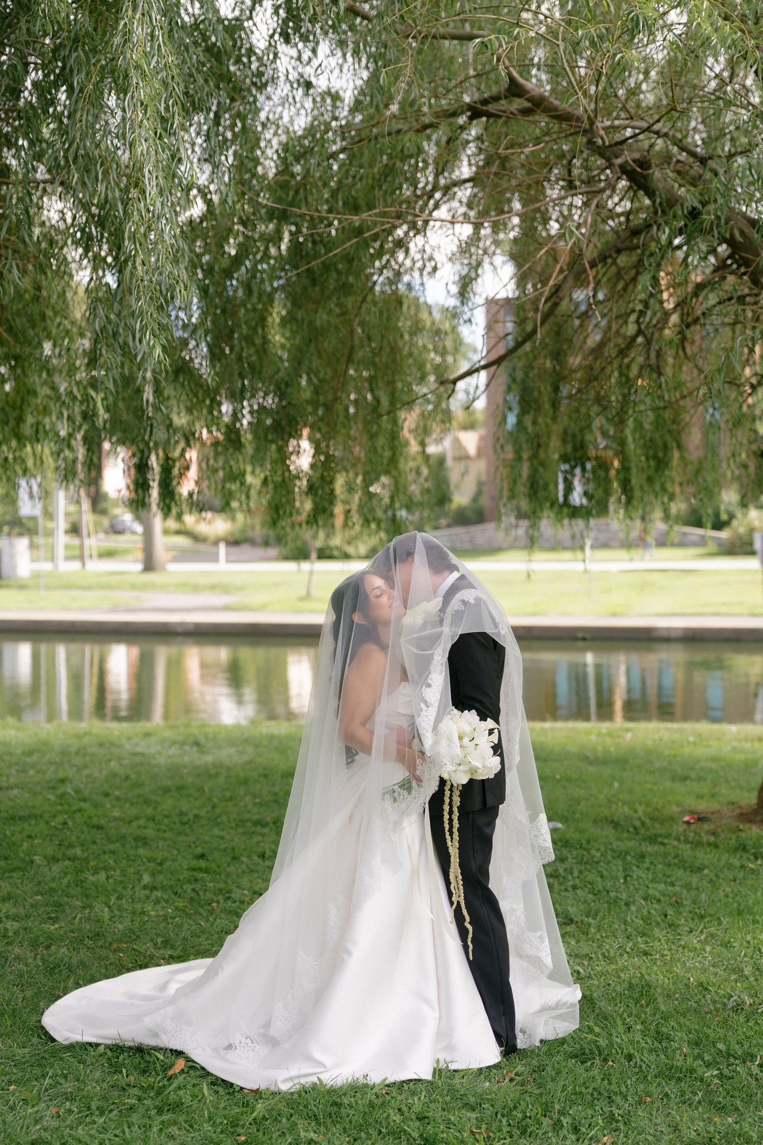 Bride and groom kissing under a flowing wedding veil by a serene pond, surrounded by weeping willow trees.