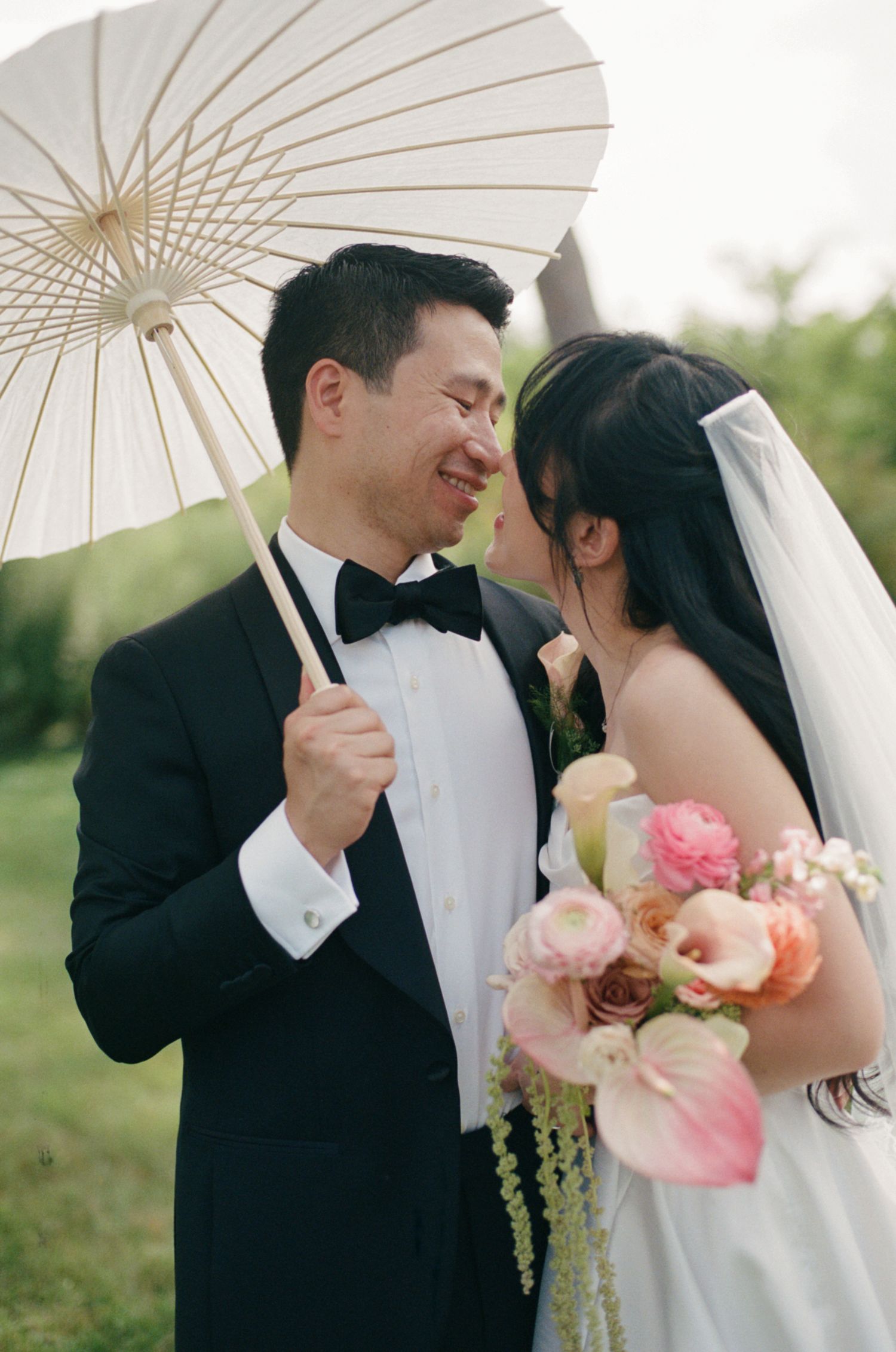 Bride and groom share a romantic moment under a white parasol, bride holding a pink floral bouquet outdoors.
