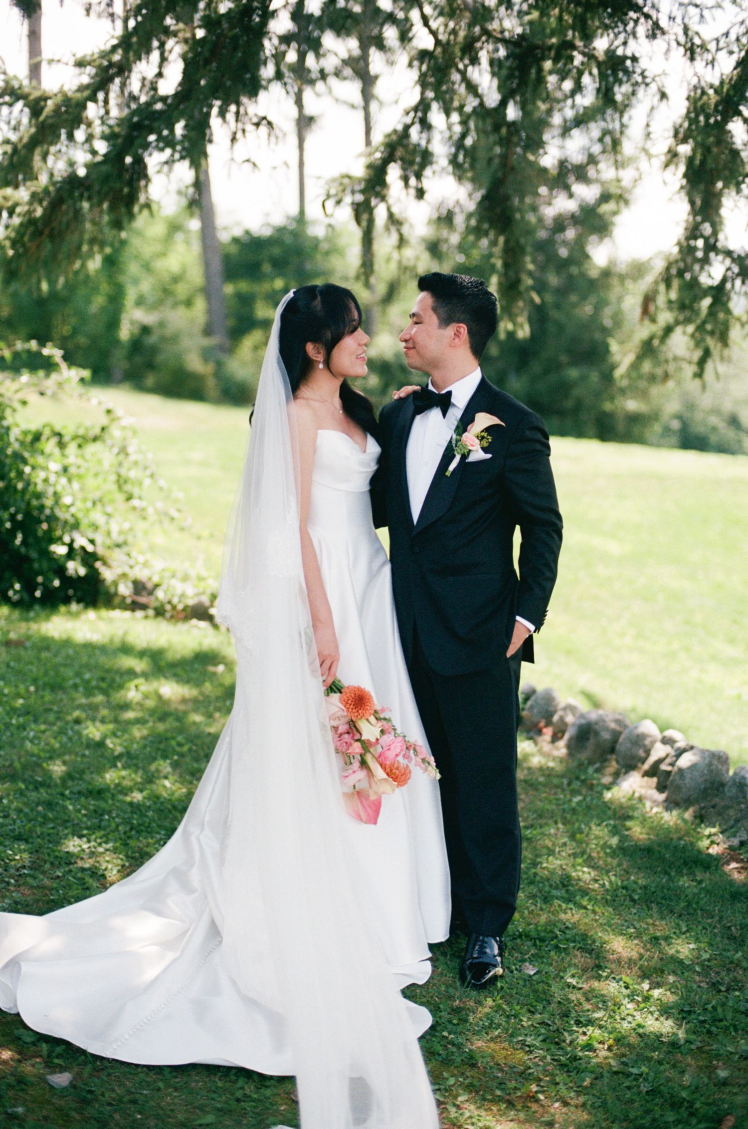 Bride in white gown with veil and groom in black tuxedo share a romantic moment outdoors on lush green lawn.