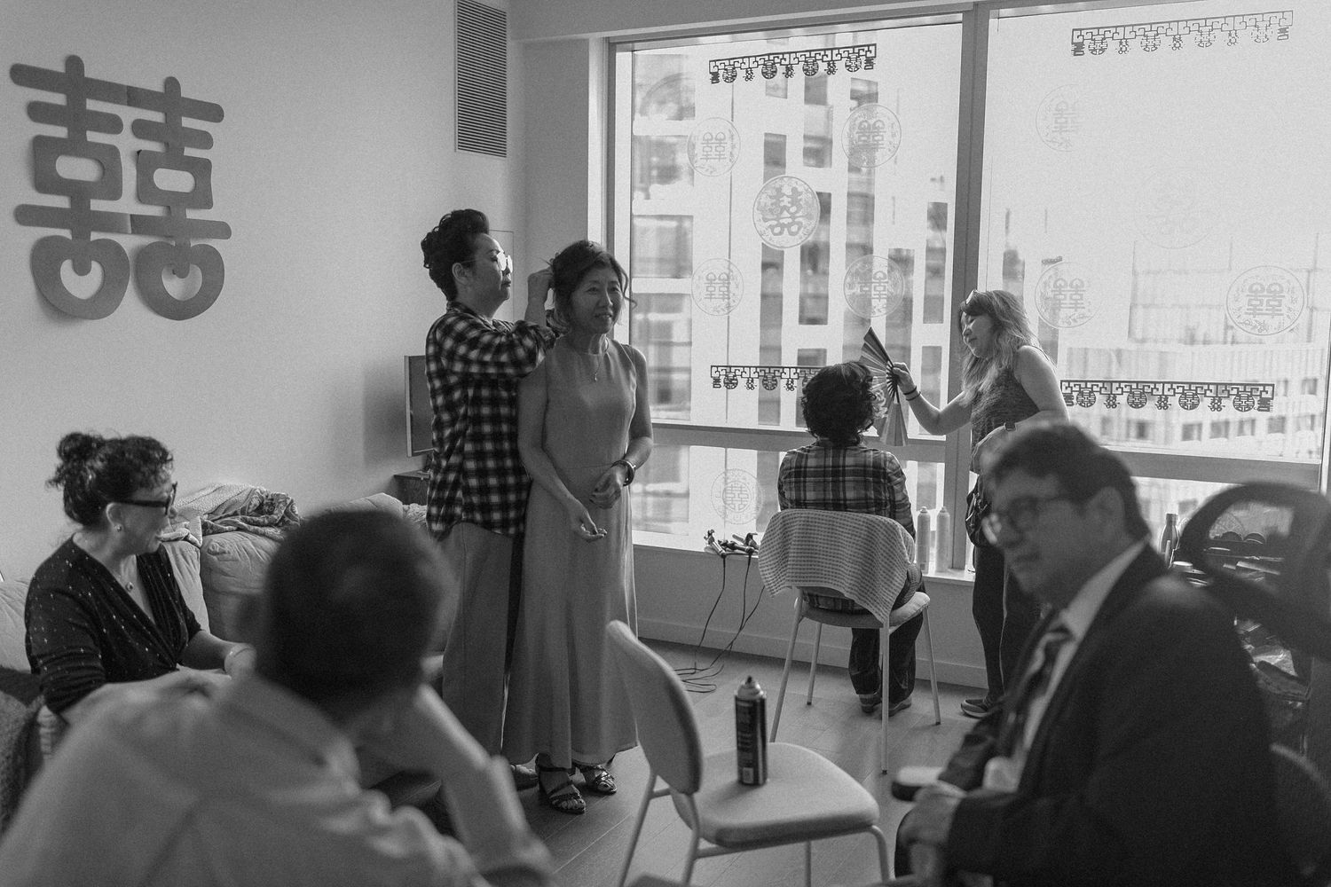 Black and white photo of a Chinese wedding preparation with guests and bridal party near a window with double happiness symbol.