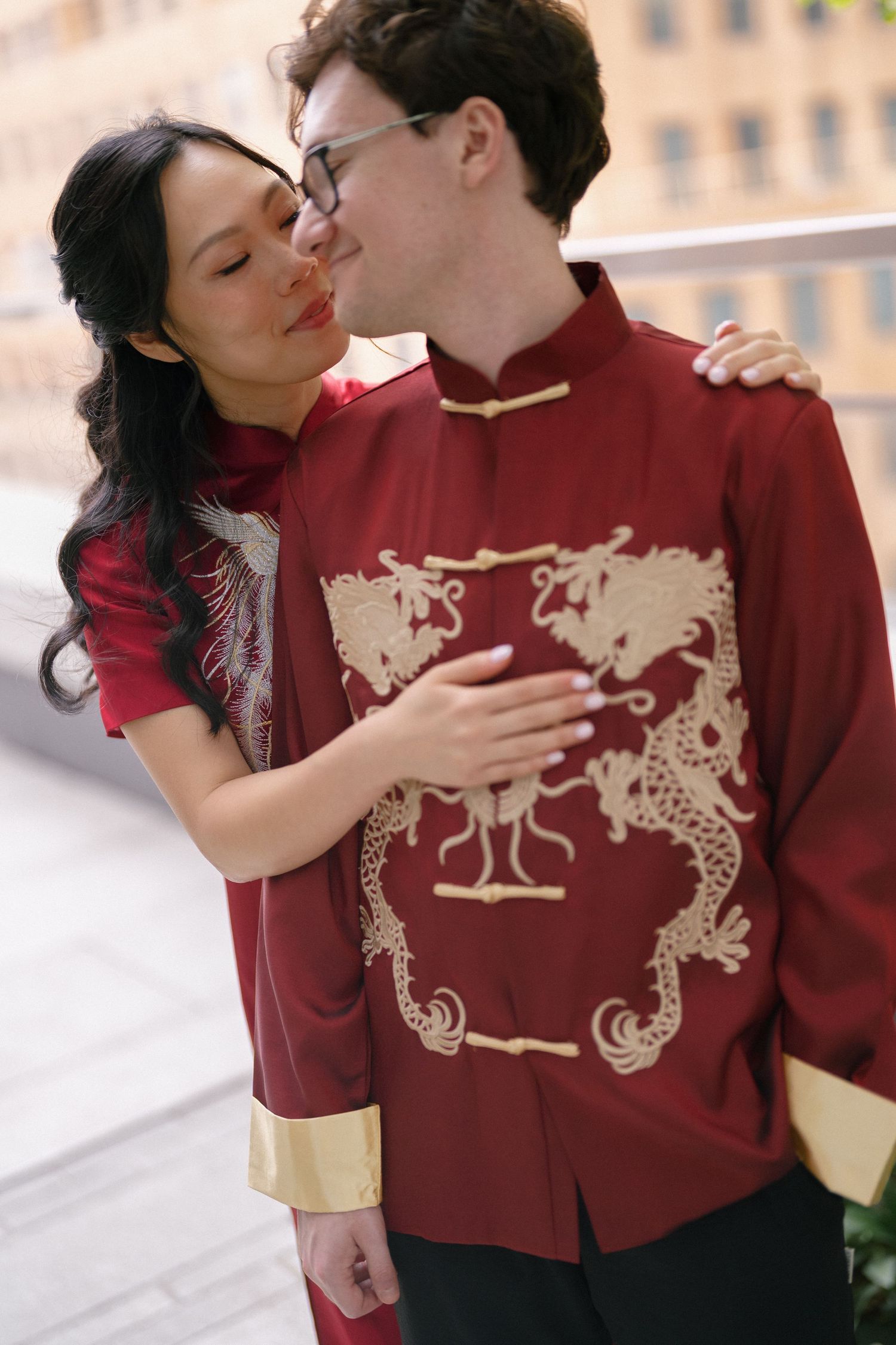 Couple in matching red Chinese traditional attire with gold dragon embroidery share a romantic moment outdoors.