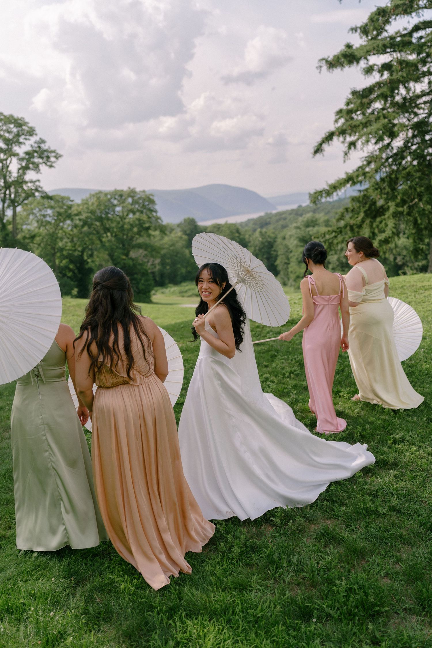 Bride in white gown poses with bridesmaids in colorful dresses holding white parasols on hillside.