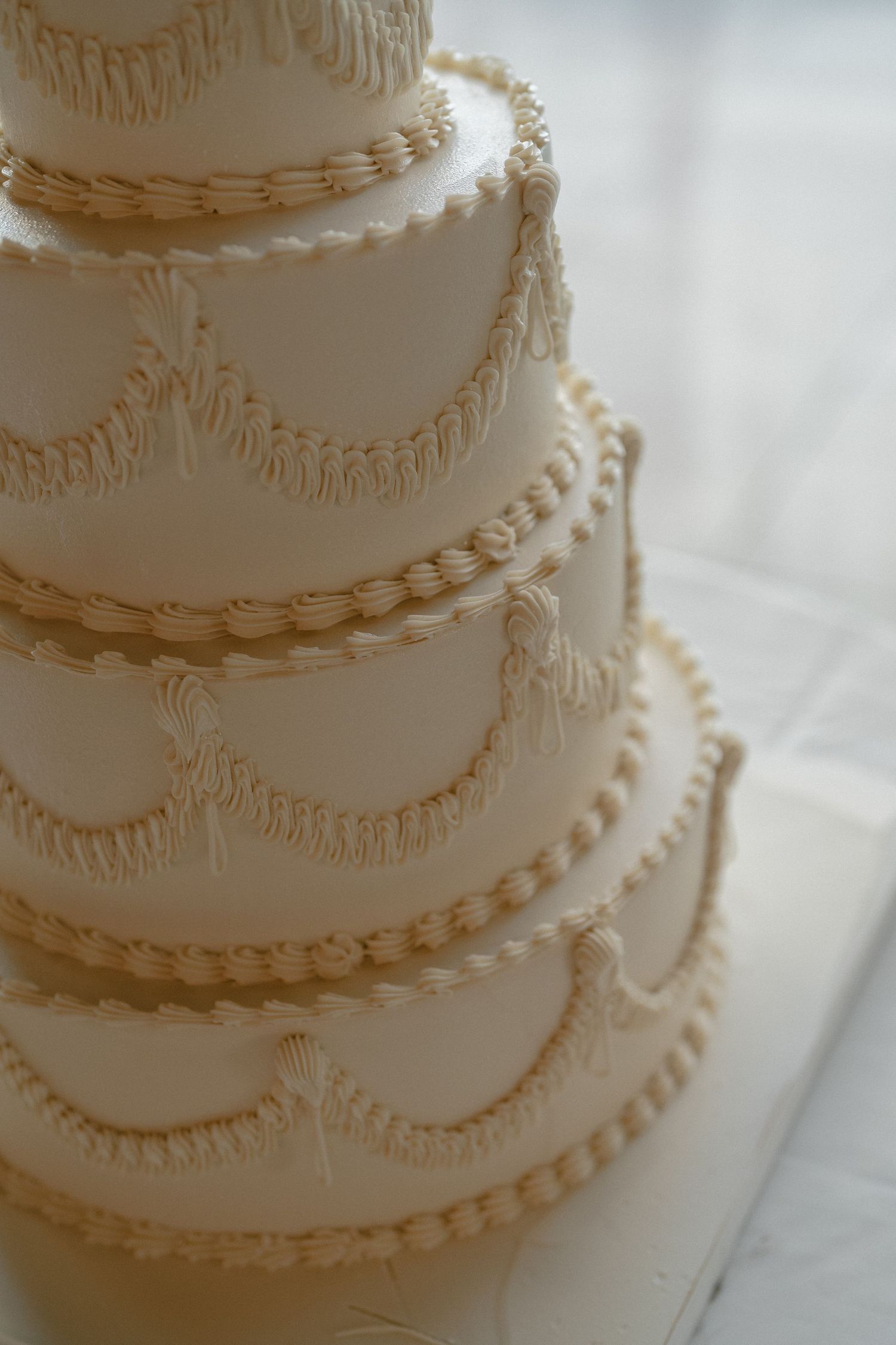 A four-tiered white wedding cake decorated with elegant piped buttercream swags, pearls, and fringe detailing.