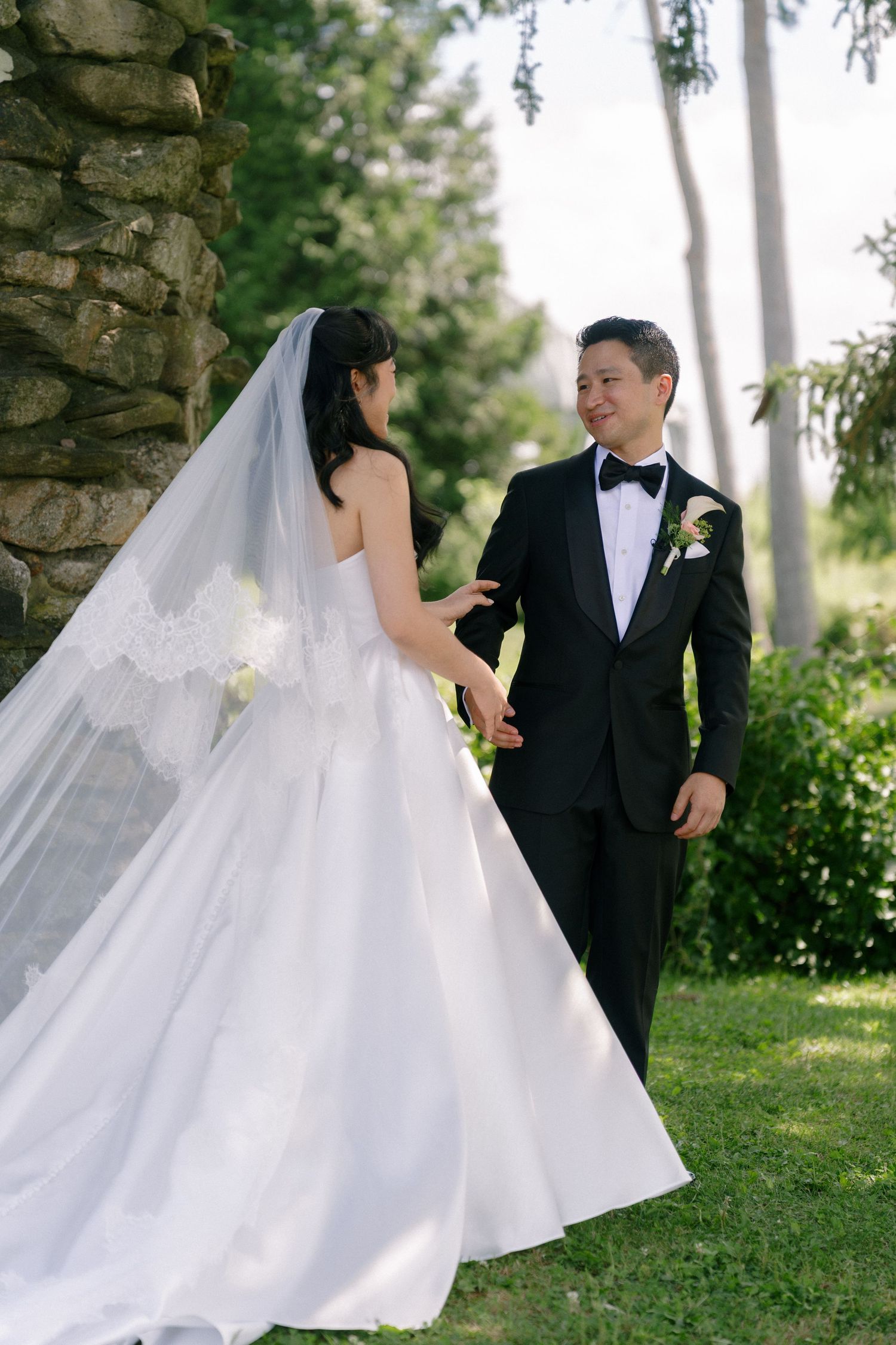 Bride in lace-trimmed veil and gown holds groom's hand as he smiles in black tuxedo outdoors.