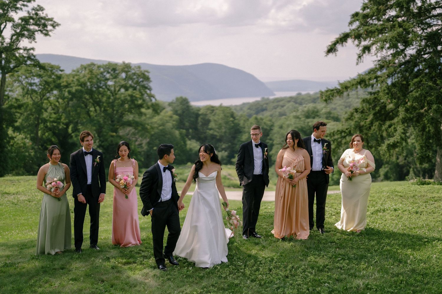 Wedding party poses on lush green lawn with scenic mountain and river views in background.