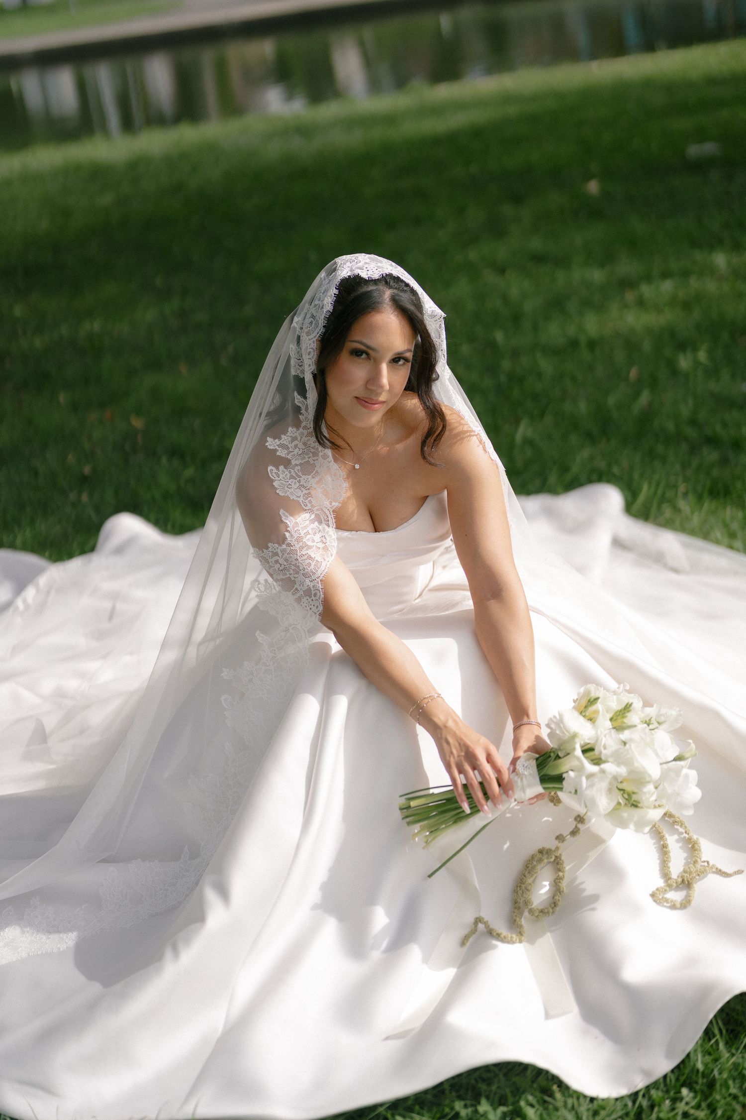 A bride in a strapless white ball gown and lace veil sits on green grass holding a white calla lily bouquet outdoors.