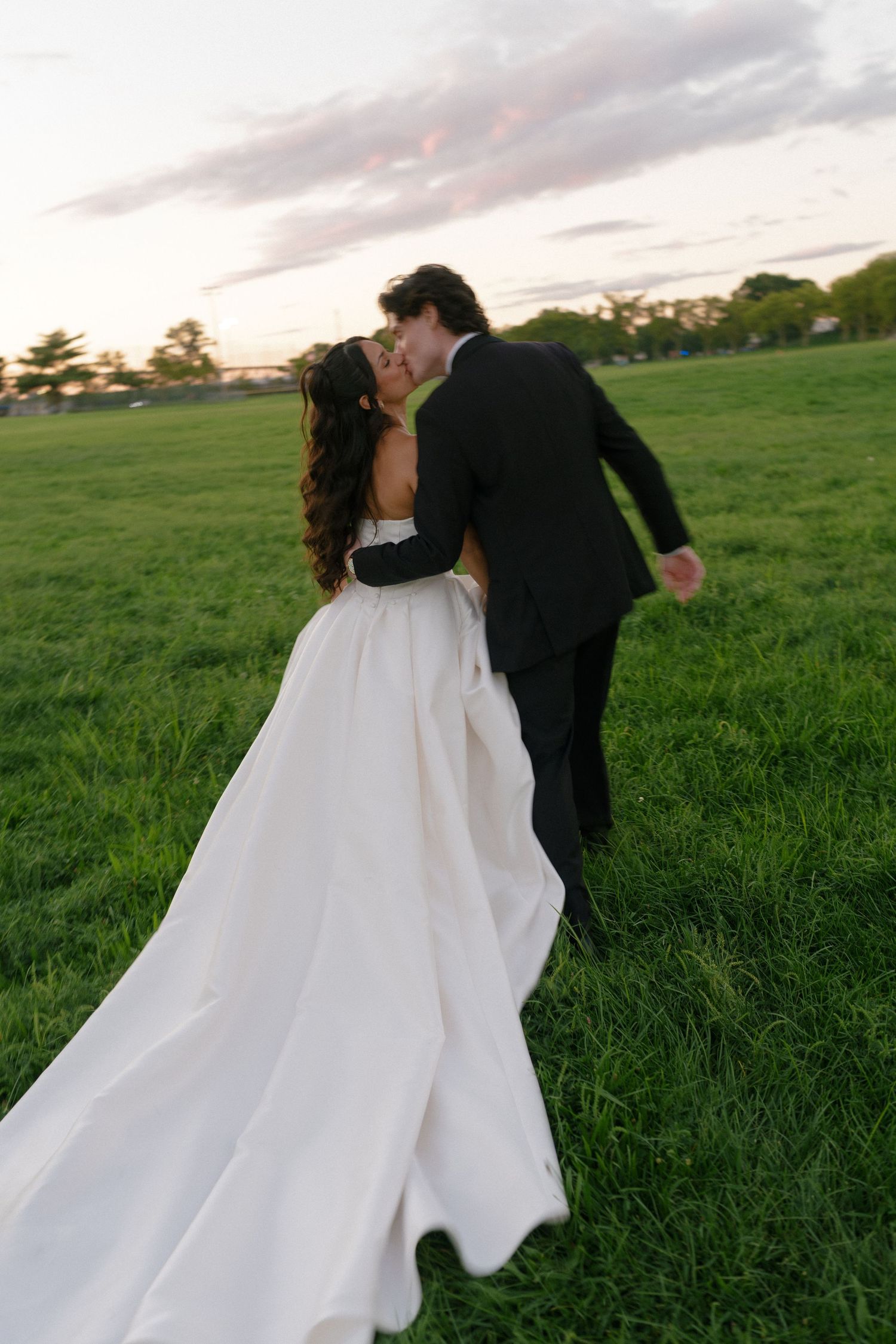Bride and groom sharing a romantic kiss in a lush green field at sunset.