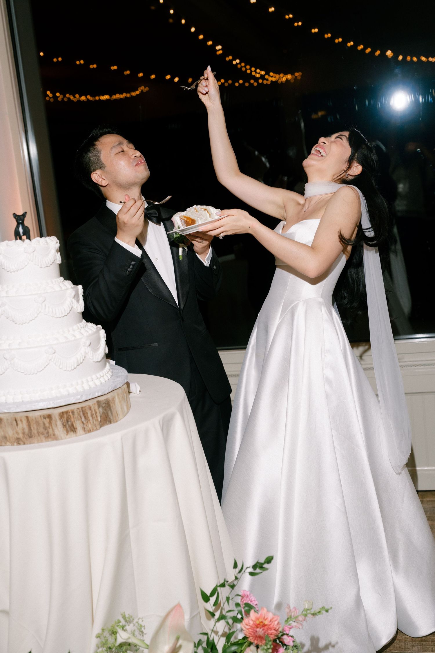 Bride and groom laugh during wedding cake cutting ceremony, bride holding cake up high at elegant evening reception.