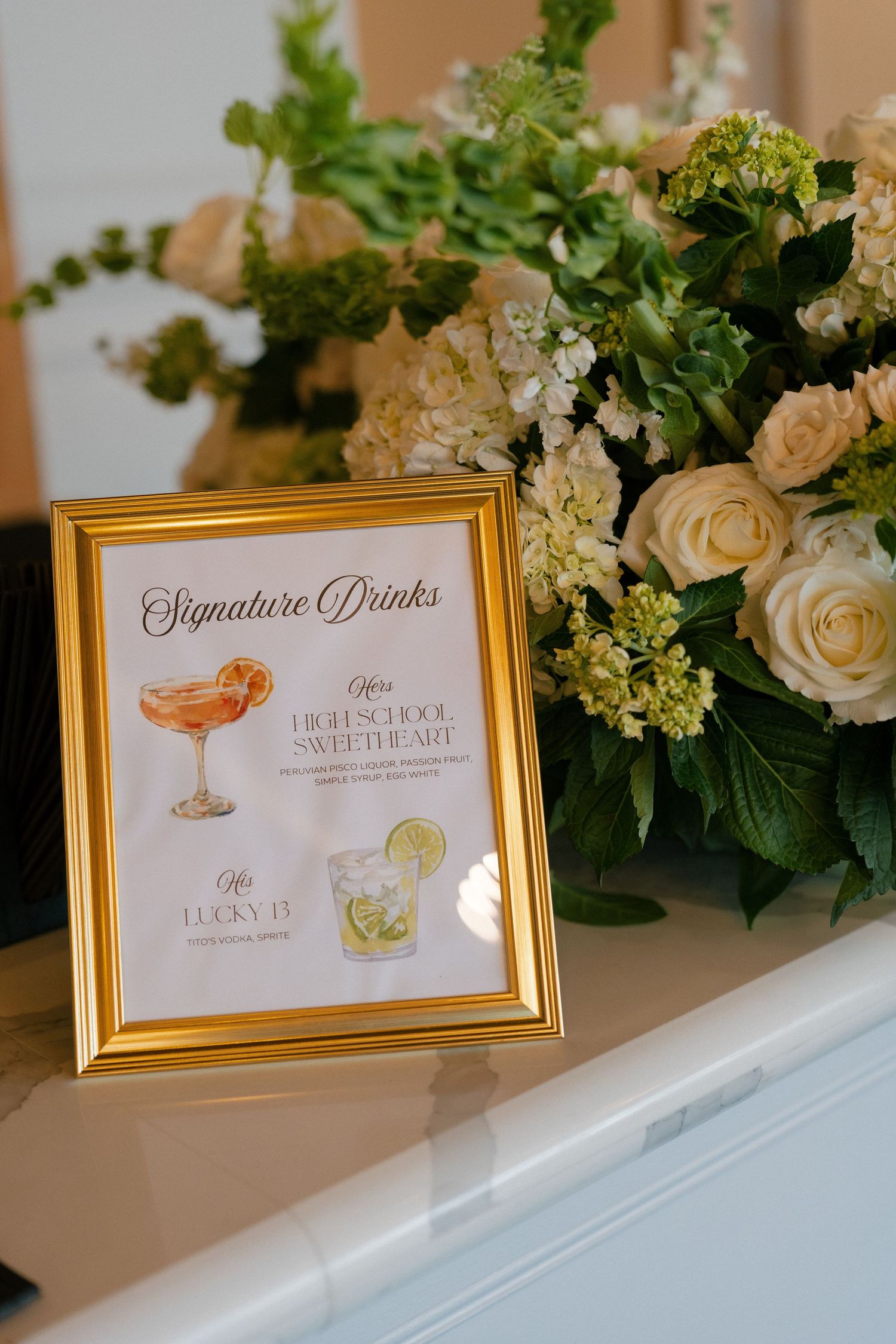 A gold-framed signature drinks menu displaying High School Sweetheart and Lucky 13 cocktails beside a lush cream floral arrangement.