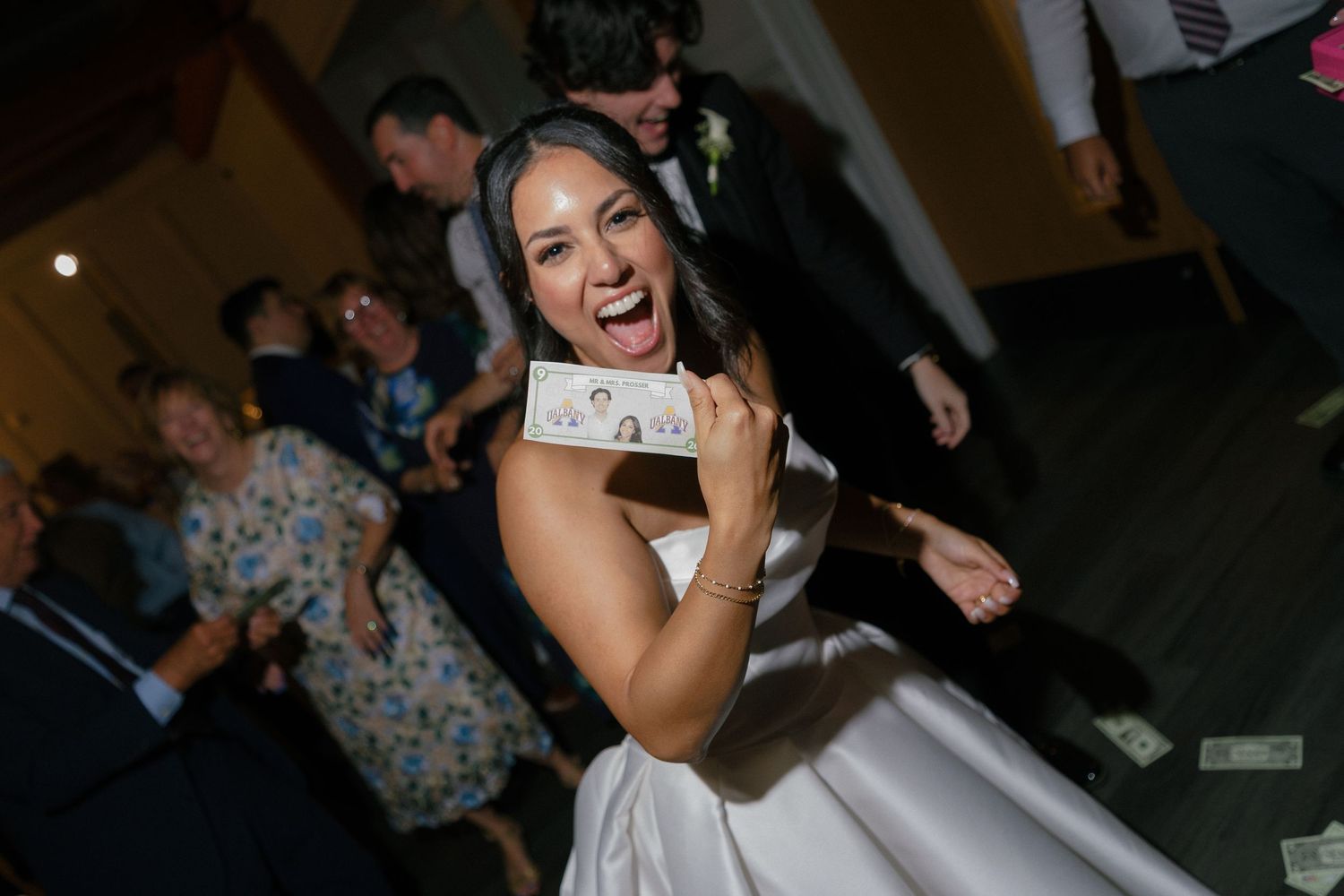 Excited bride in white gown holding cash money while dancing at wedding reception celebration.