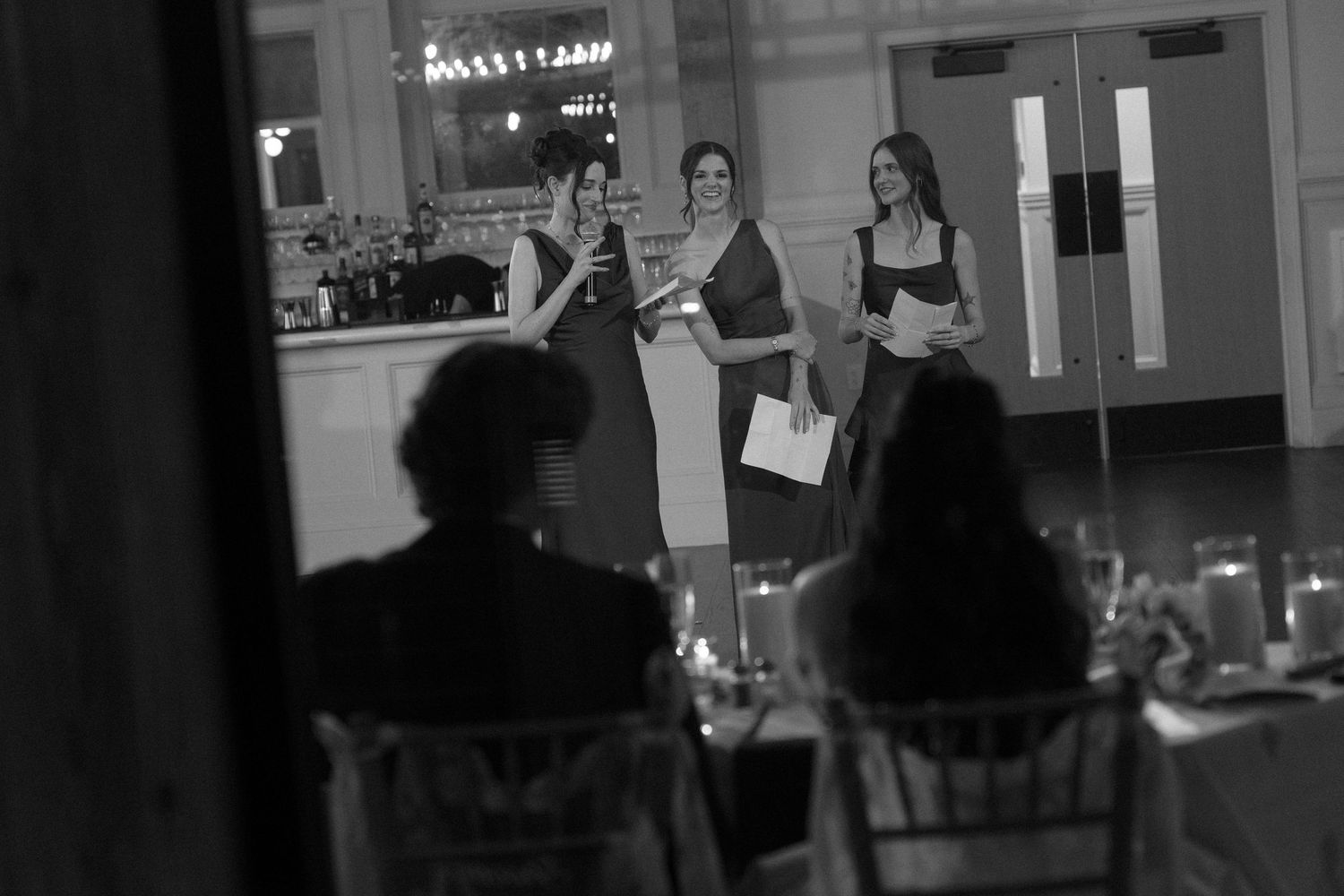 Black and white photo of three women giving toasts at elegant wedding reception while guests watch from tables.