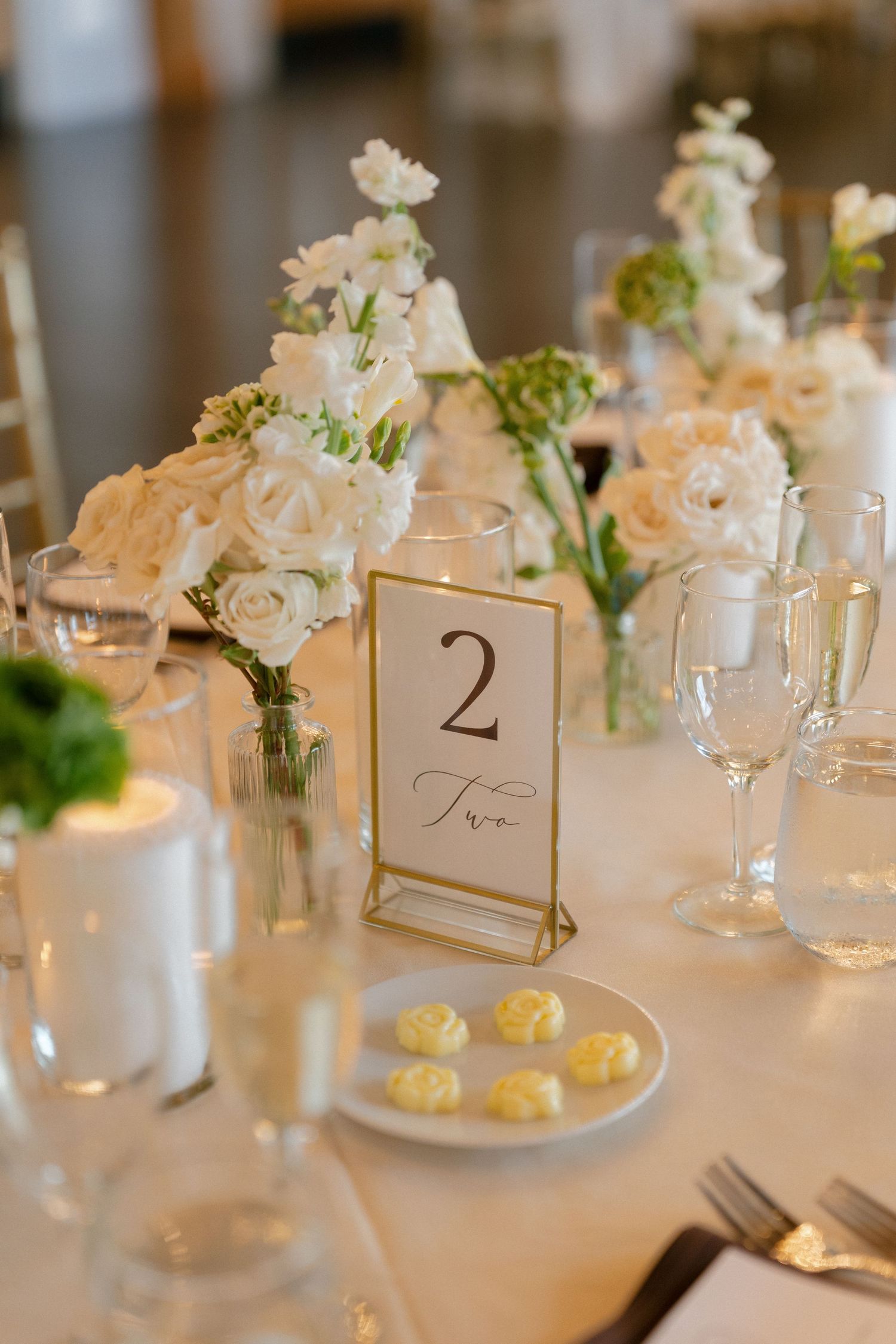 White floral centerpiece with roses beside gold-framed table number 2 at wedding reception.