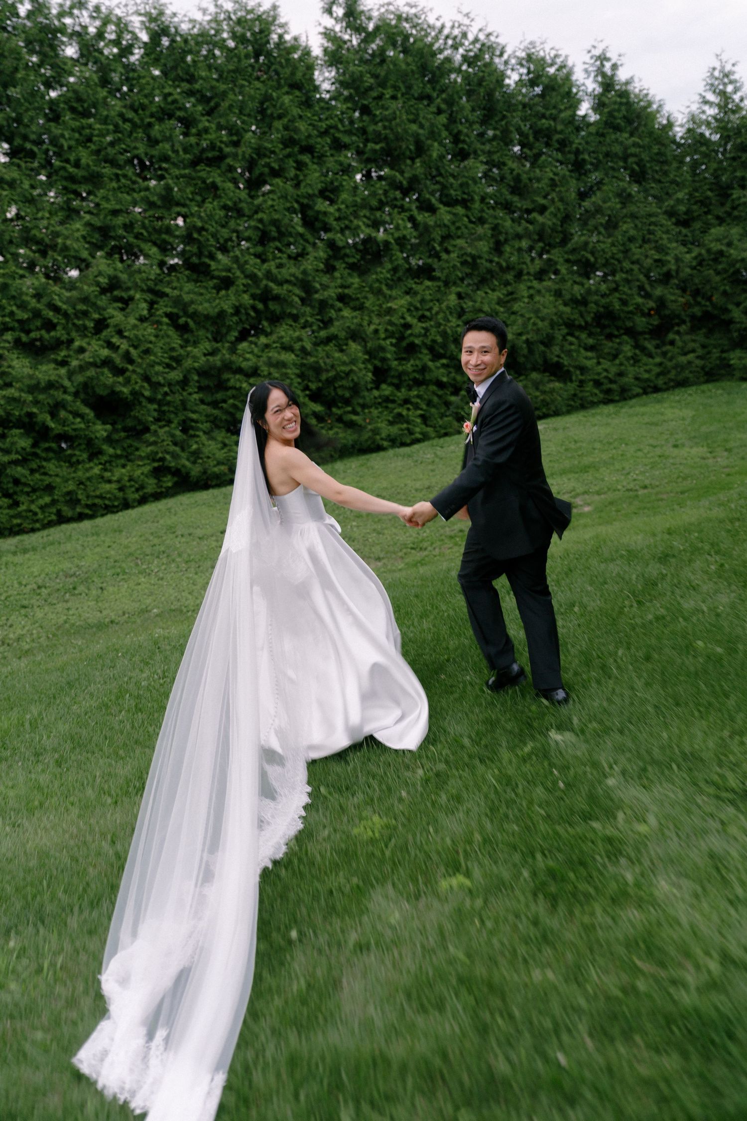 Bride and groom holding hands running on green lawn, brides long veil flowing behind her.