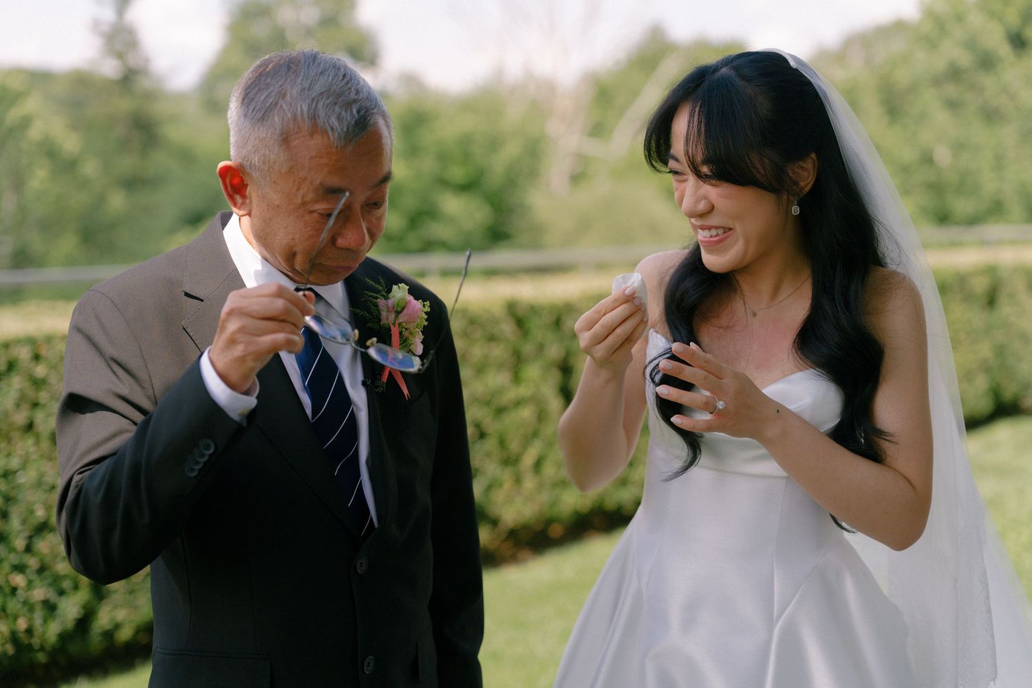 Bride in white gown and older man in suit blow bubbles together outdoors at a wedding celebration.