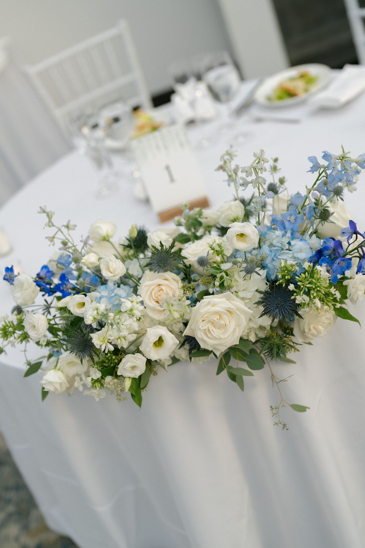 Elegant wedding table centerpiece with white roses and blue delphinium flowers on a white tablecloth.