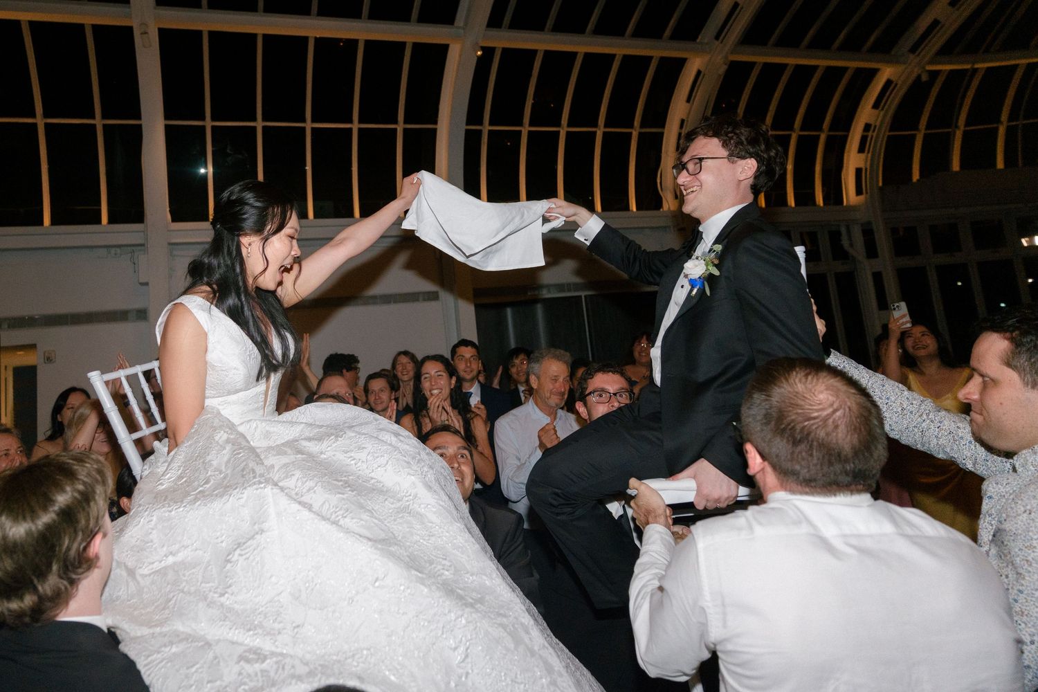 Bride and groom lifted by guests during joyful Jewish wedding hora dance celebration at elegant indoor venue.