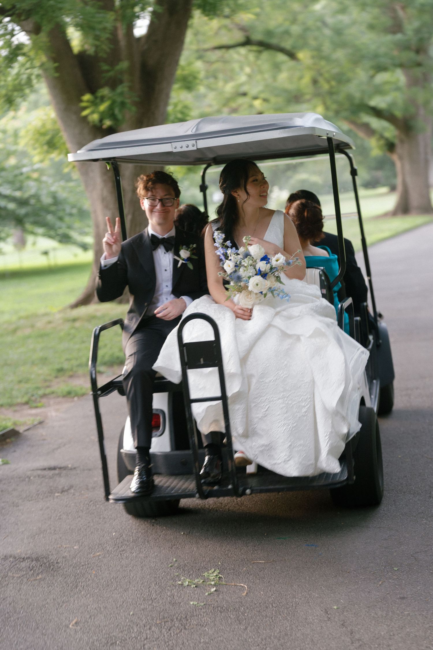 Bride and groom riding a golf cart on their wedding day, groom flashing peace sign, bride holding bouquet.