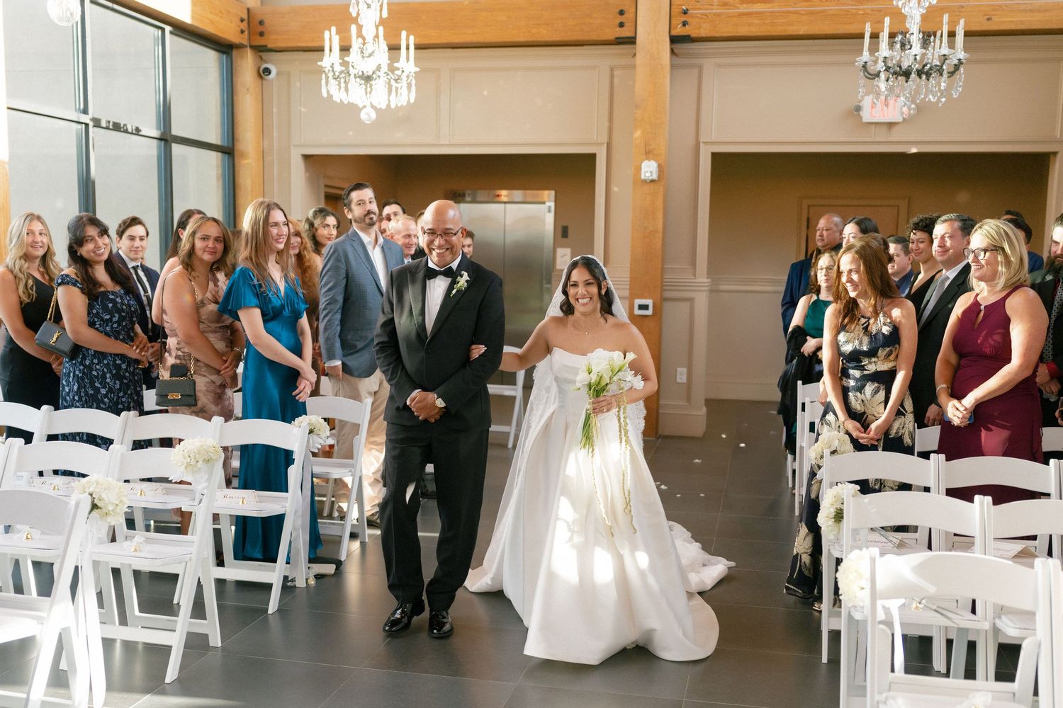 Bride walks down the aisle with her father in a elegant wedding venue with white chairs and chandelier lighting.