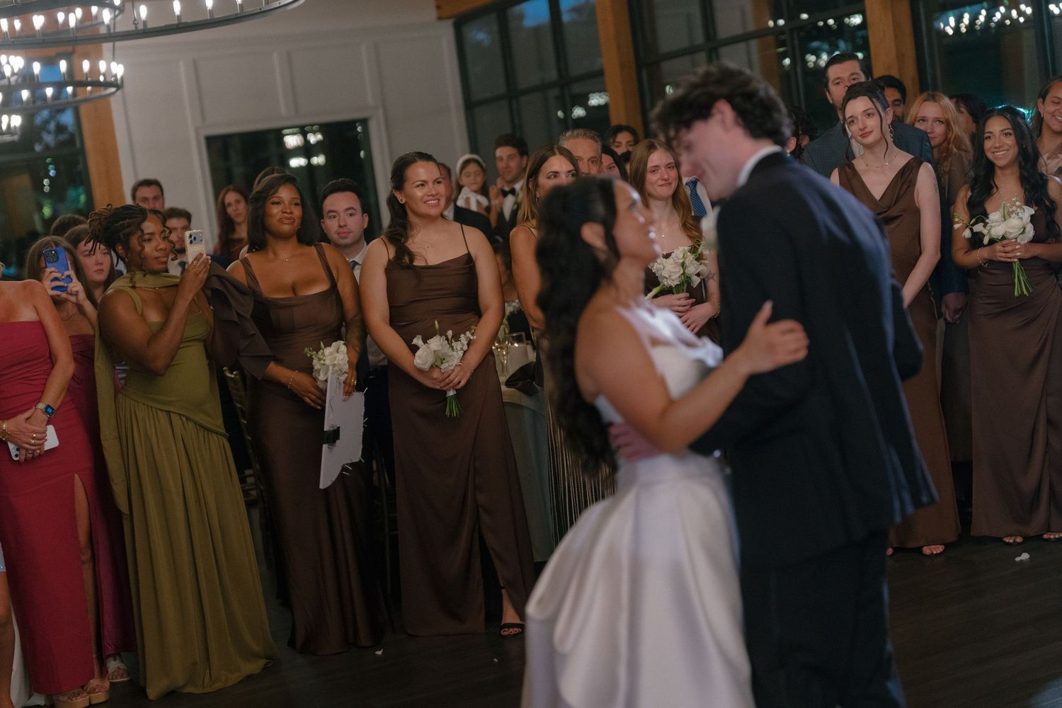 Bride and groom share first dance while bridesmaids and guests look on smiling.