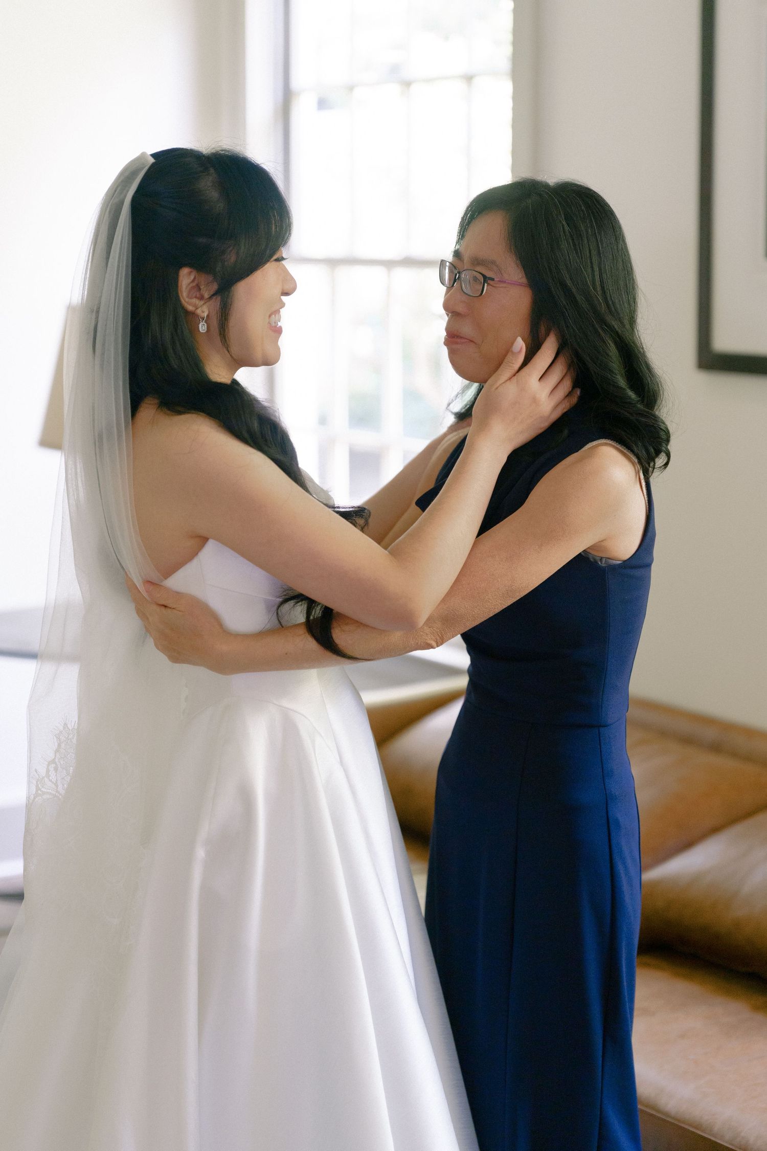 Bride in white gown and veil shares tender moment with mother in navy dress before wedding ceremony.