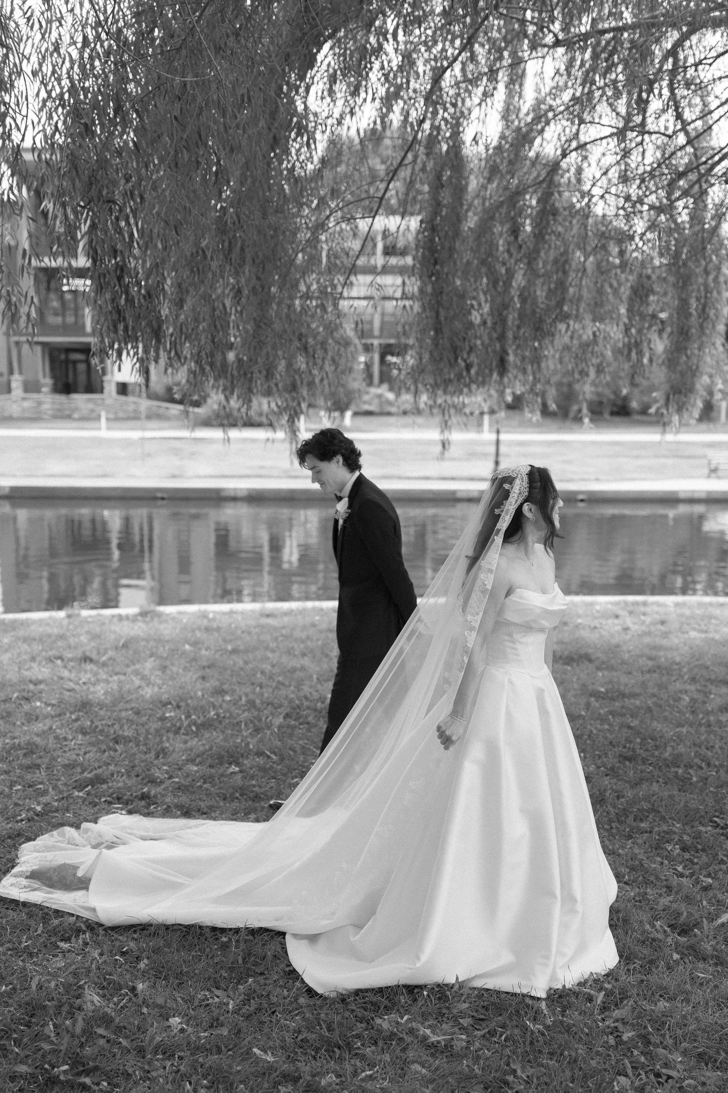 Black and white photo of a bride and groom standing by a pond under a weeping willow tree.