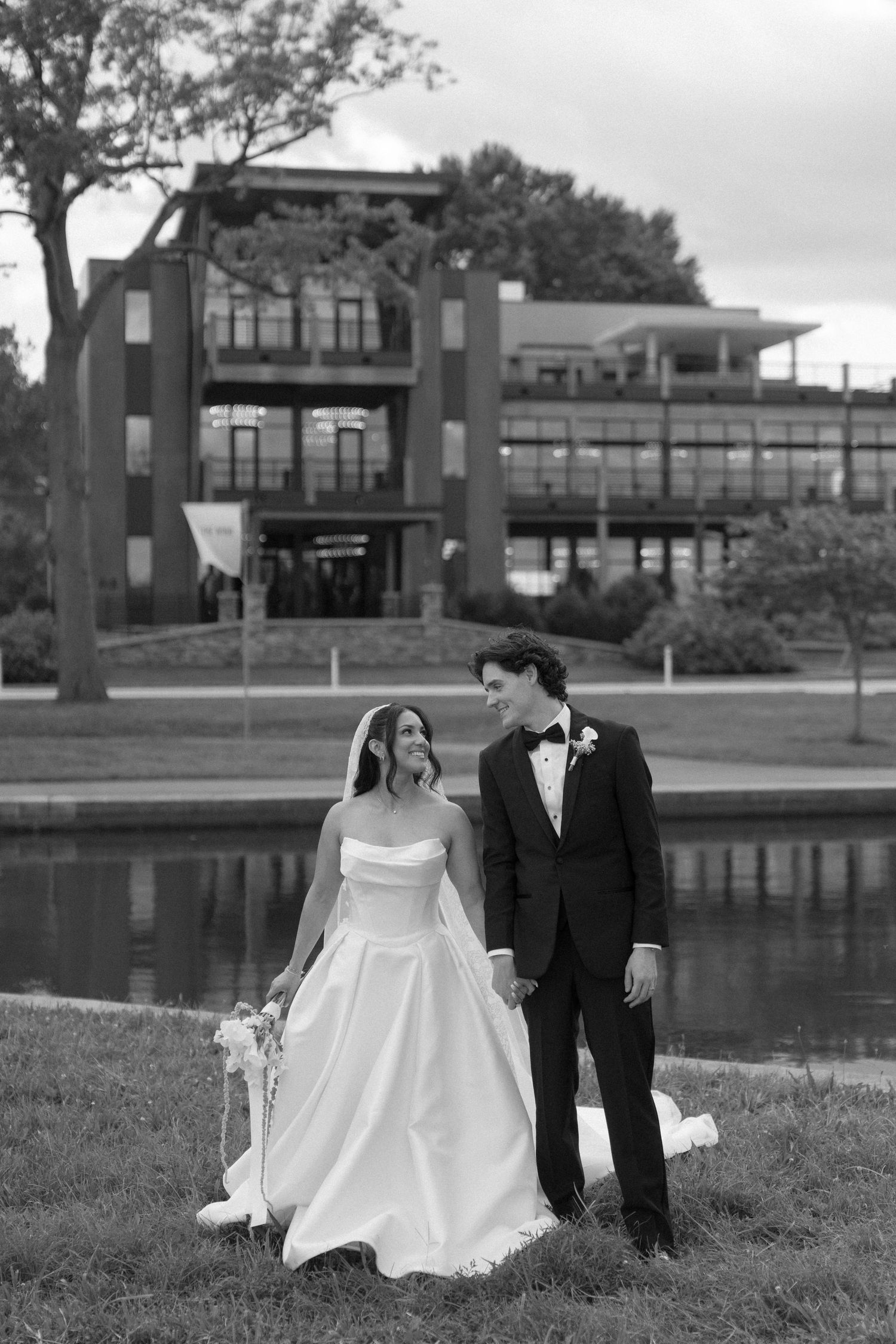 Black and white photo of bride and groom holding hands walking by a pond with a building backdrop.