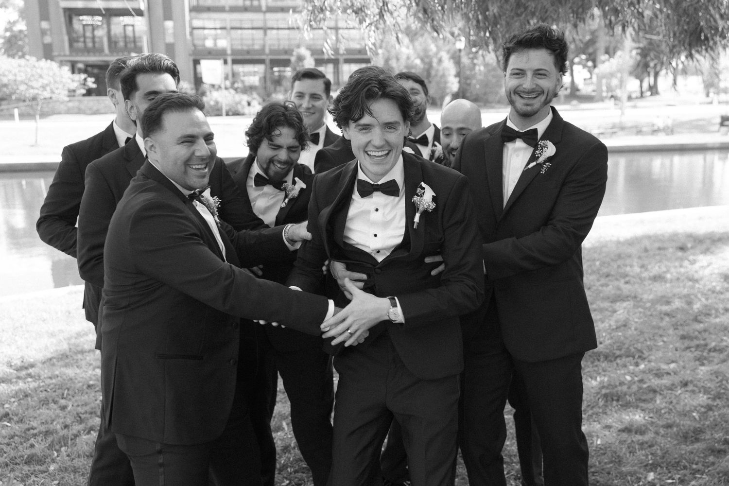 Groom surrounded by groomsmen in black tuxedos laughing outdoors in black and white wedding photo.