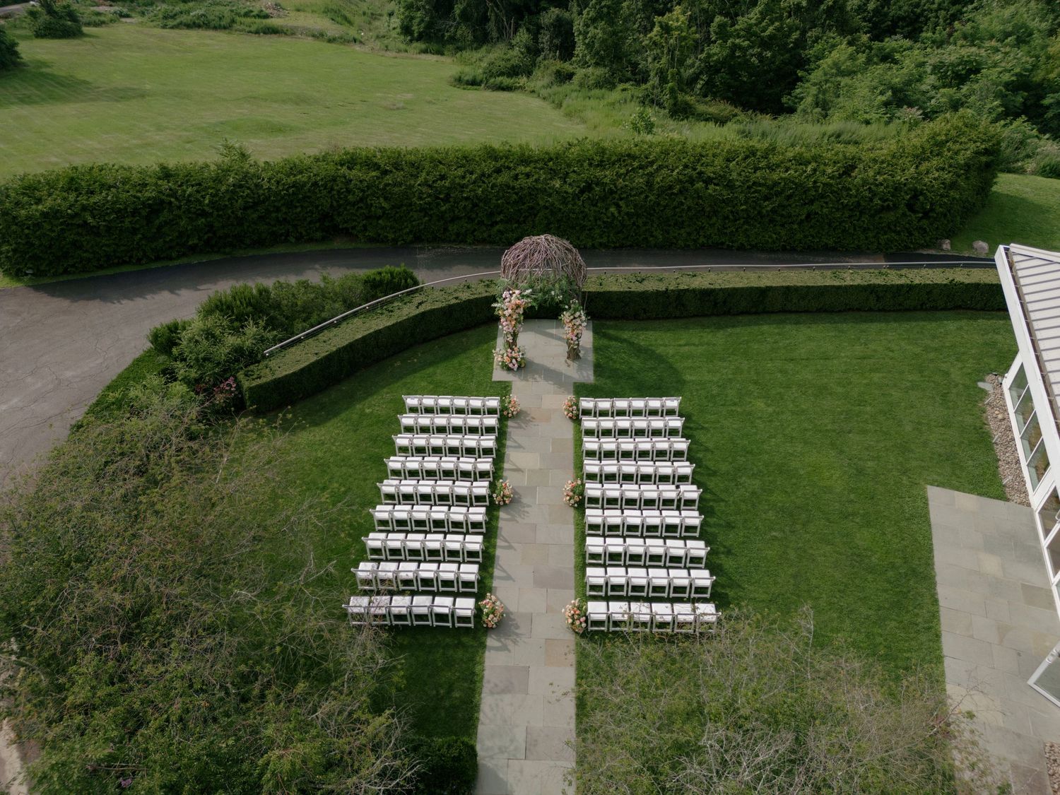Aerial view of an outdoor wedding ceremony setup with white chairs arranged in rows on a green lawn with a floral arch.
