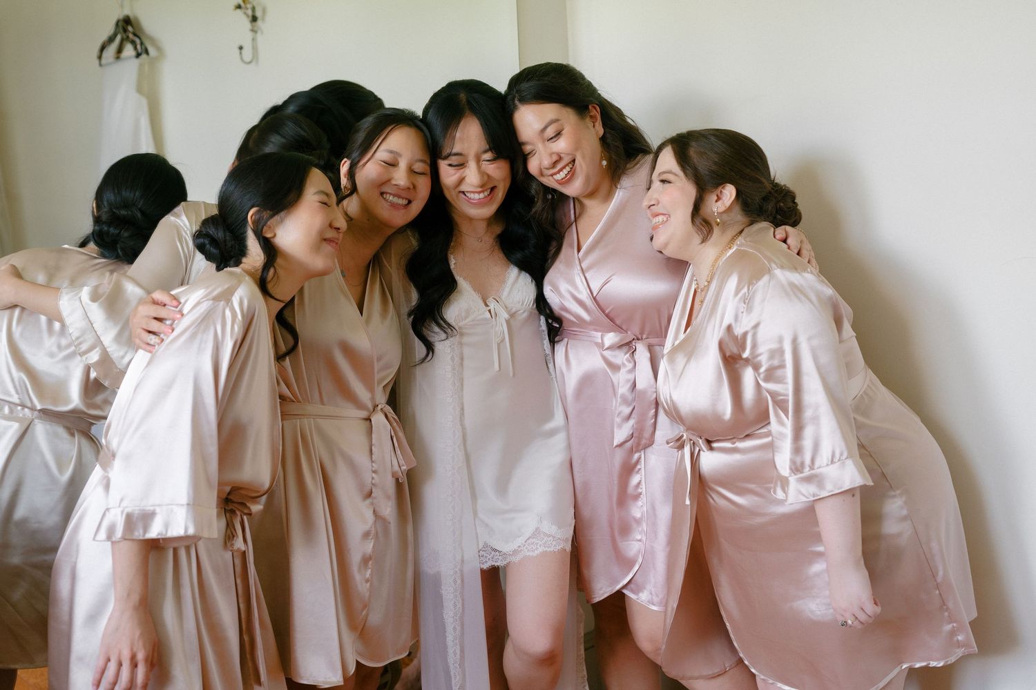 Bridesmaids in blush satin robes laughing and hugging with bride in white dress during wedding morning prep.