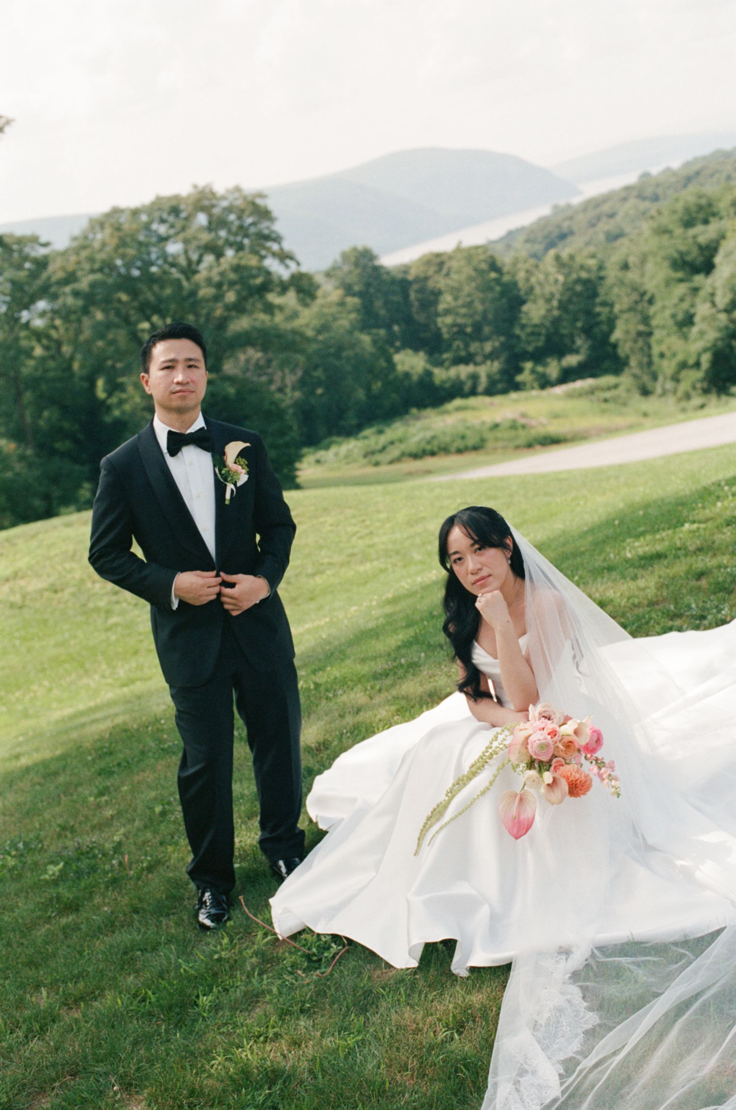 Bride sitting on green hillside holding pink bouquet with groom standing beside her, mountains behind.