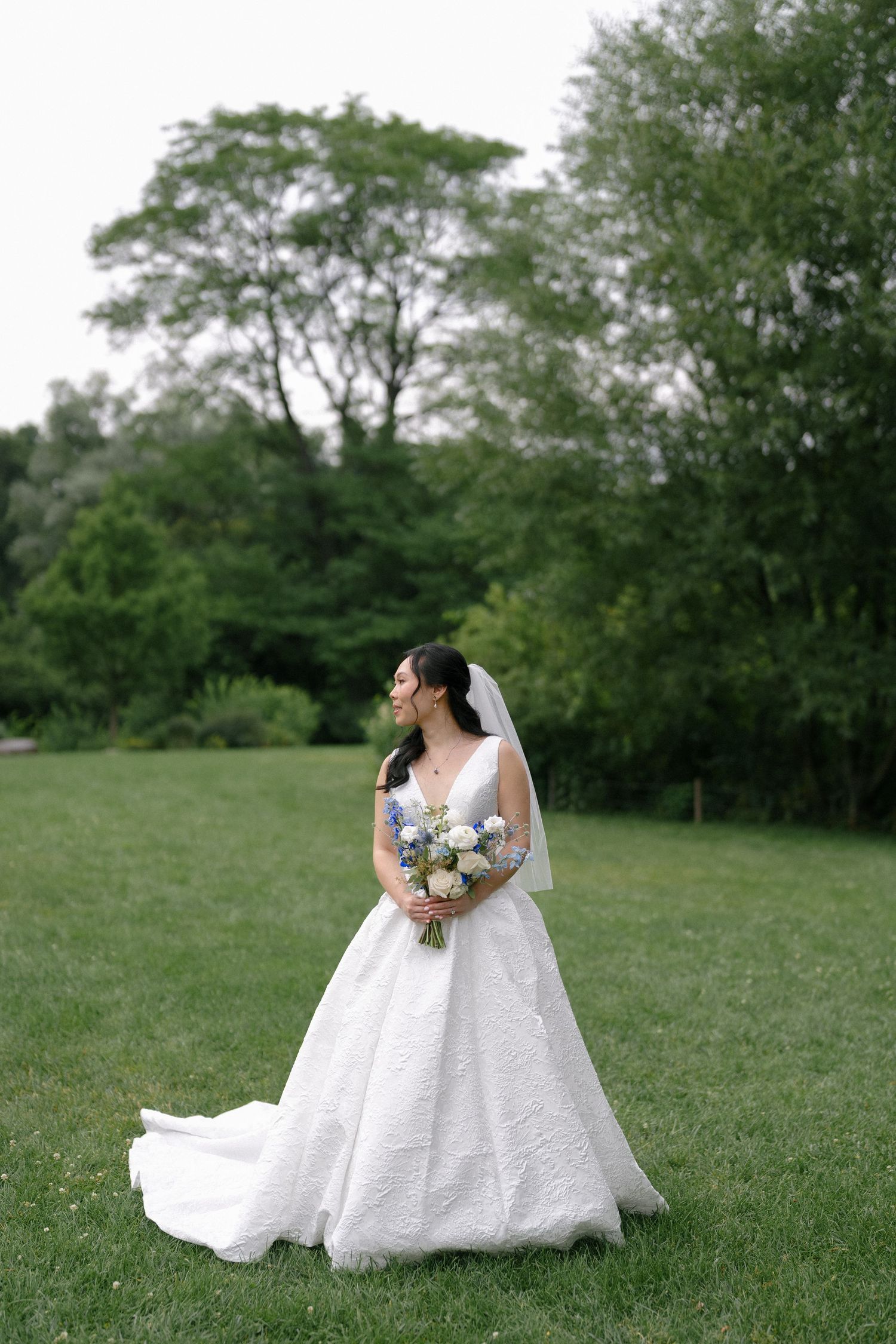 Elegant bride in white ballgown with veil holds floral bouquet while standing on lush green lawn.