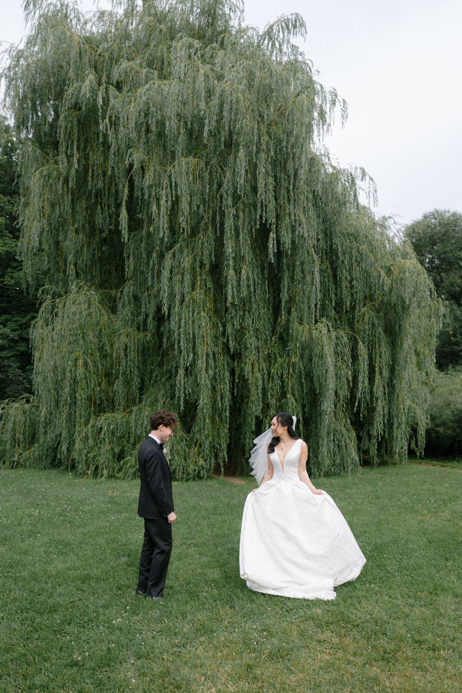 Bride and groom share a romantic first look beneath a weeping willow tree on lush green grass.