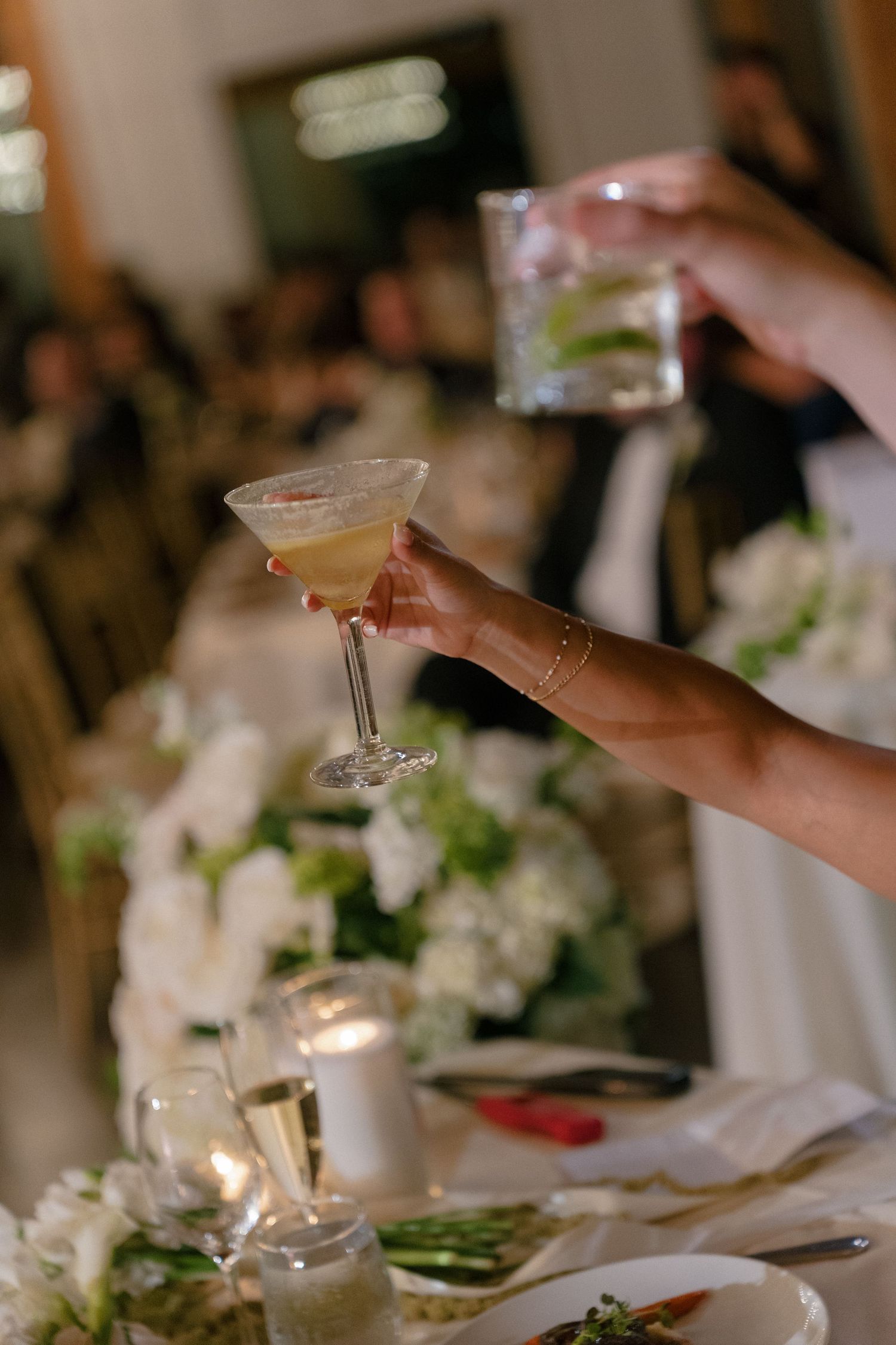 Hands clinking cocktail glasses over white floral centerpieces at elegant wedding reception dinner.