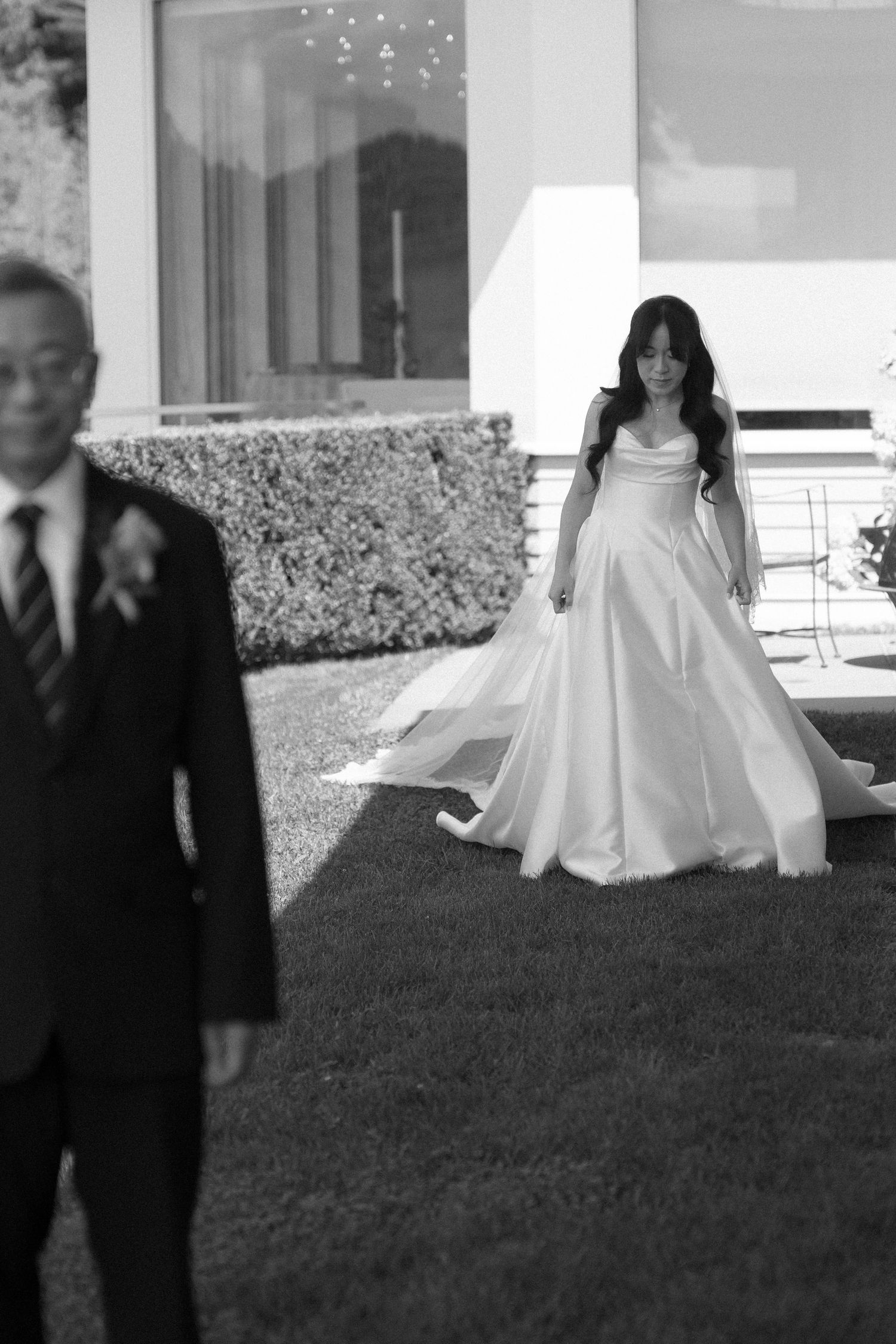Black and white photo of a bride in a flowing white gown walking toward a man in a suit on a green lawn.