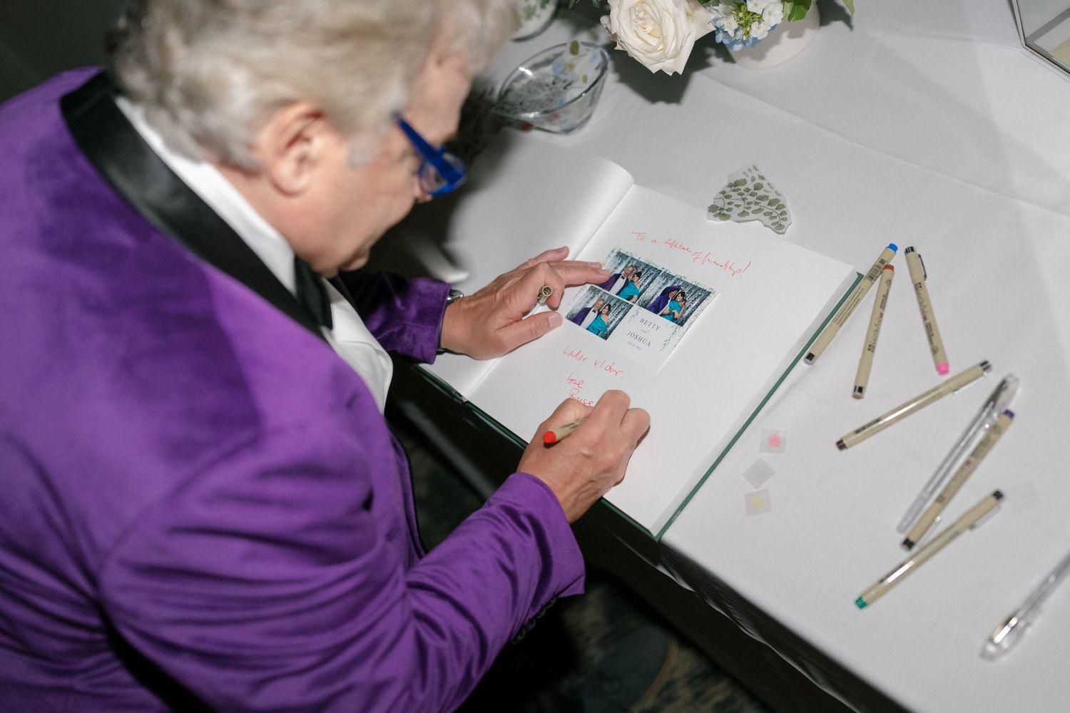 Wedding guest in purple tuxedo signs guest book with colorful markers at decorated reception table.
