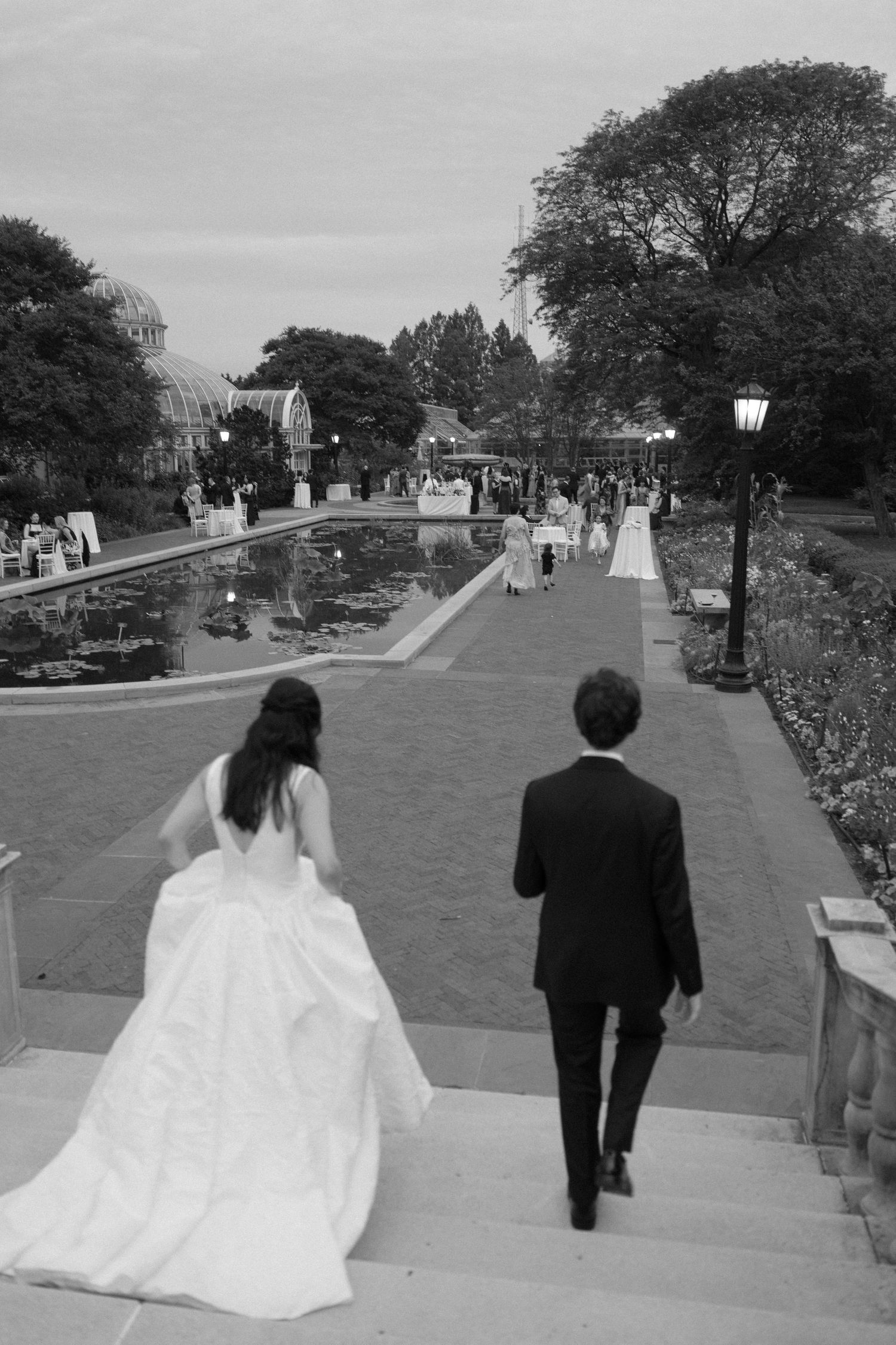 Bride and groom walk hand in hand toward outdoor wedding reception beside a lily pond garden.