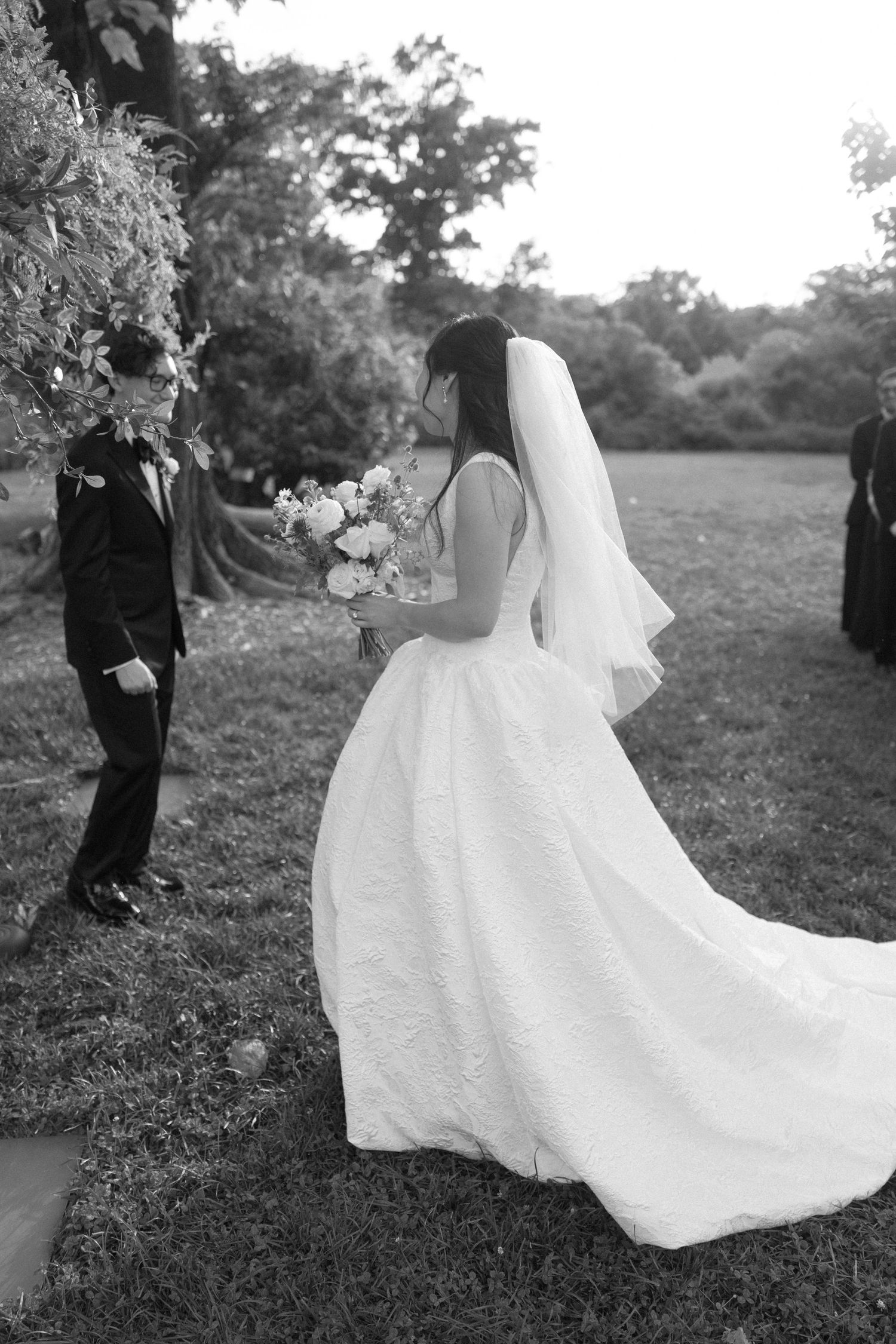 Bride in white ball gown with veil holds bouquet outdoors, facing groom in black tuxedo near floral arch.