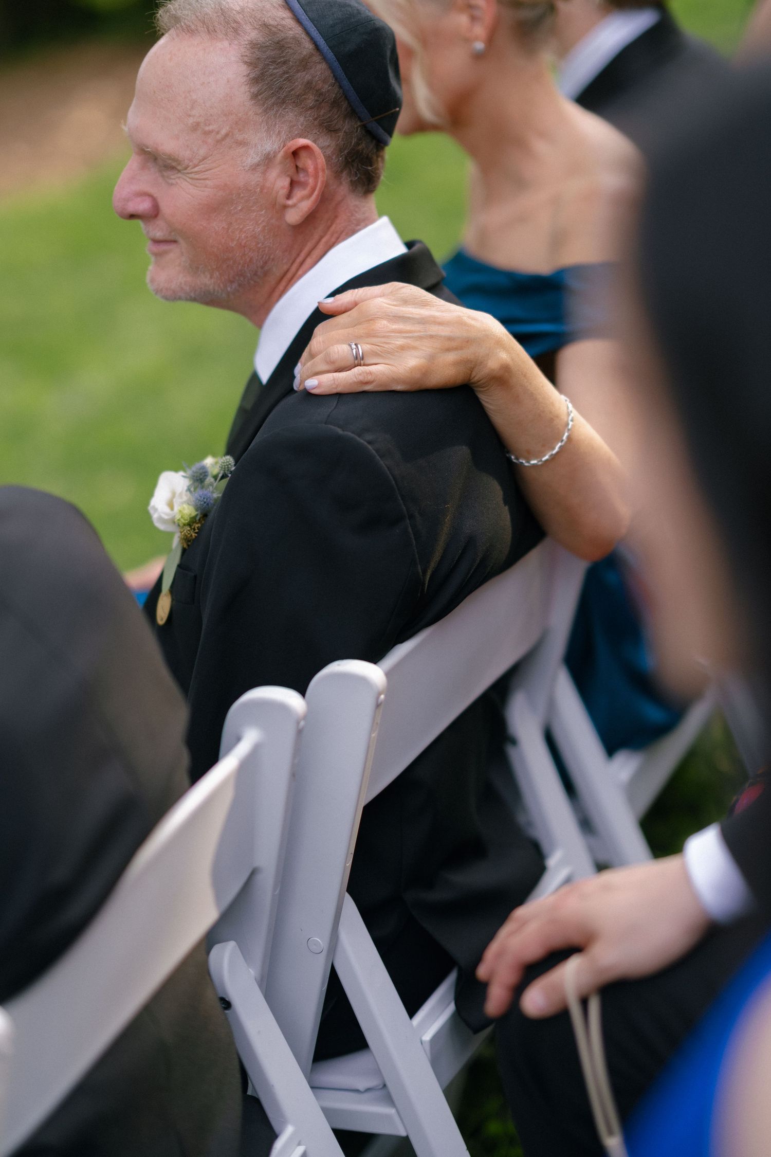 Man in black suit with yarmulke and boutonniere seated at outdoor Jewish wedding ceremony.