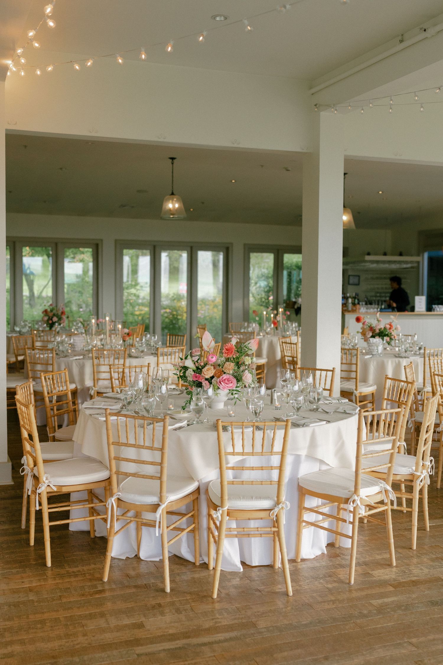 Elegant wedding reception hall with gold chiavari chairs, white tablecloths, pink floral centerpieces, and string lights.