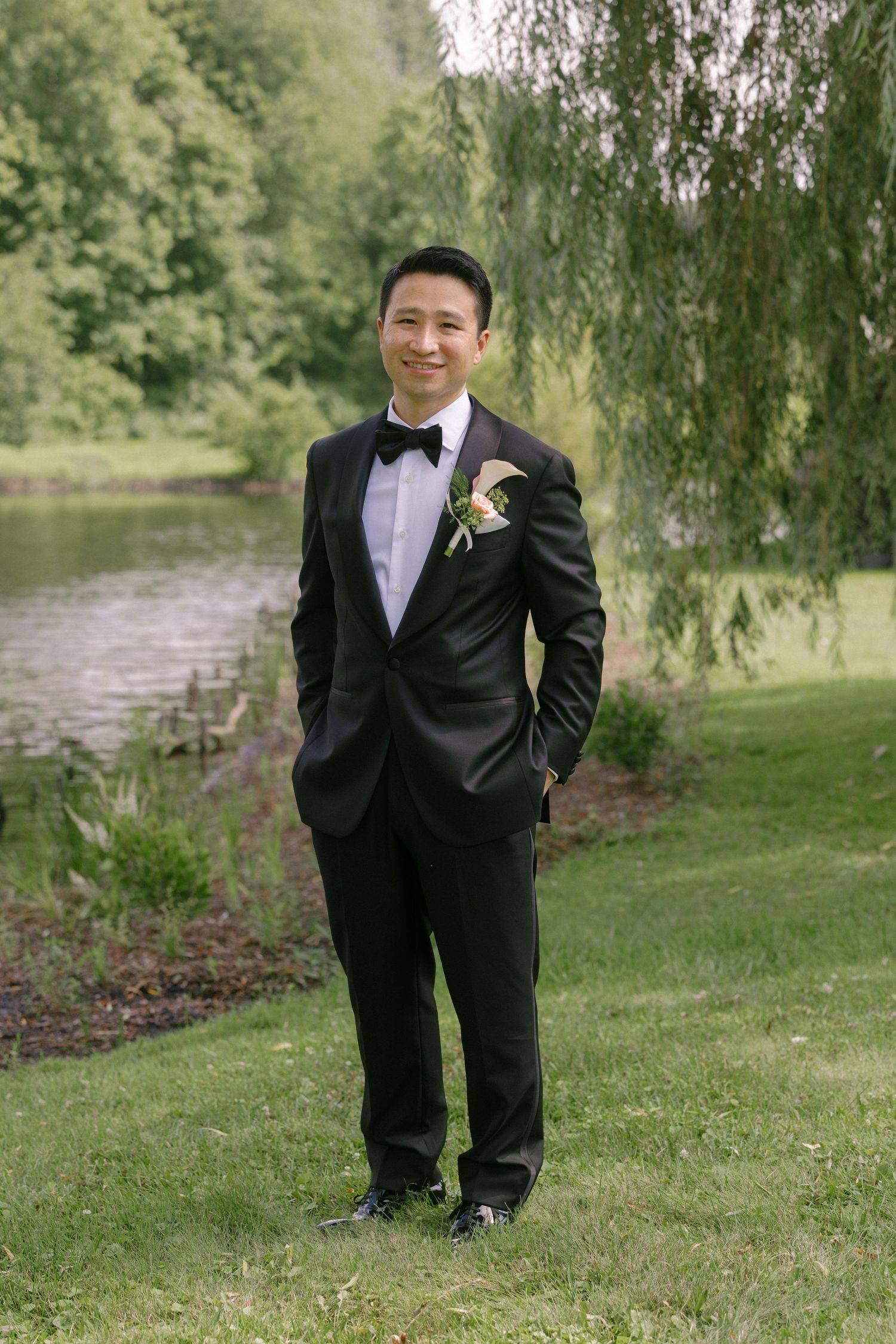 Groom in black tuxedo with boutonniere stands smiling outdoors near a pond with willow trees.